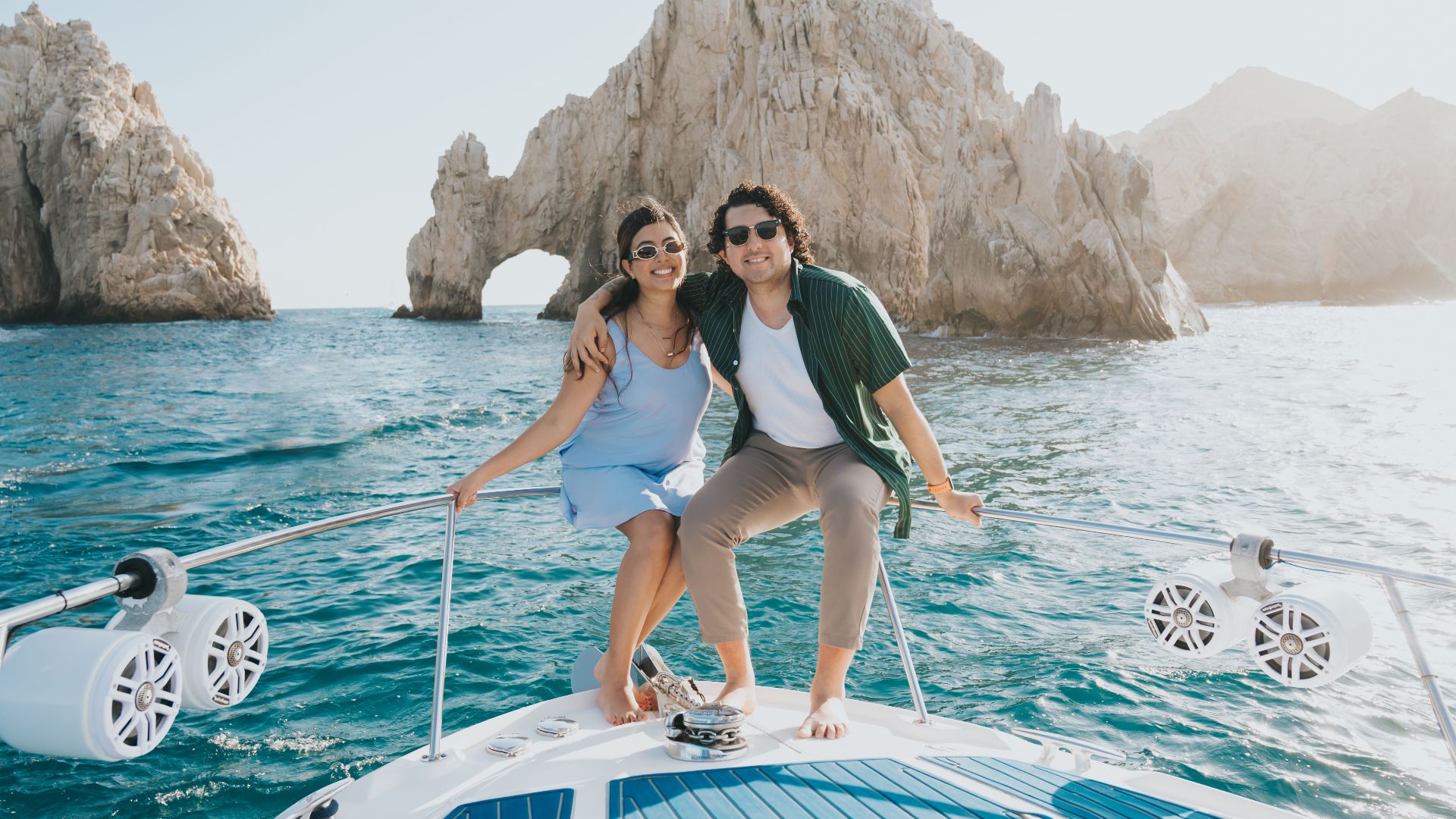 Smiling couple on boat with Arch of Cabo San Lucas in background, sunny day.