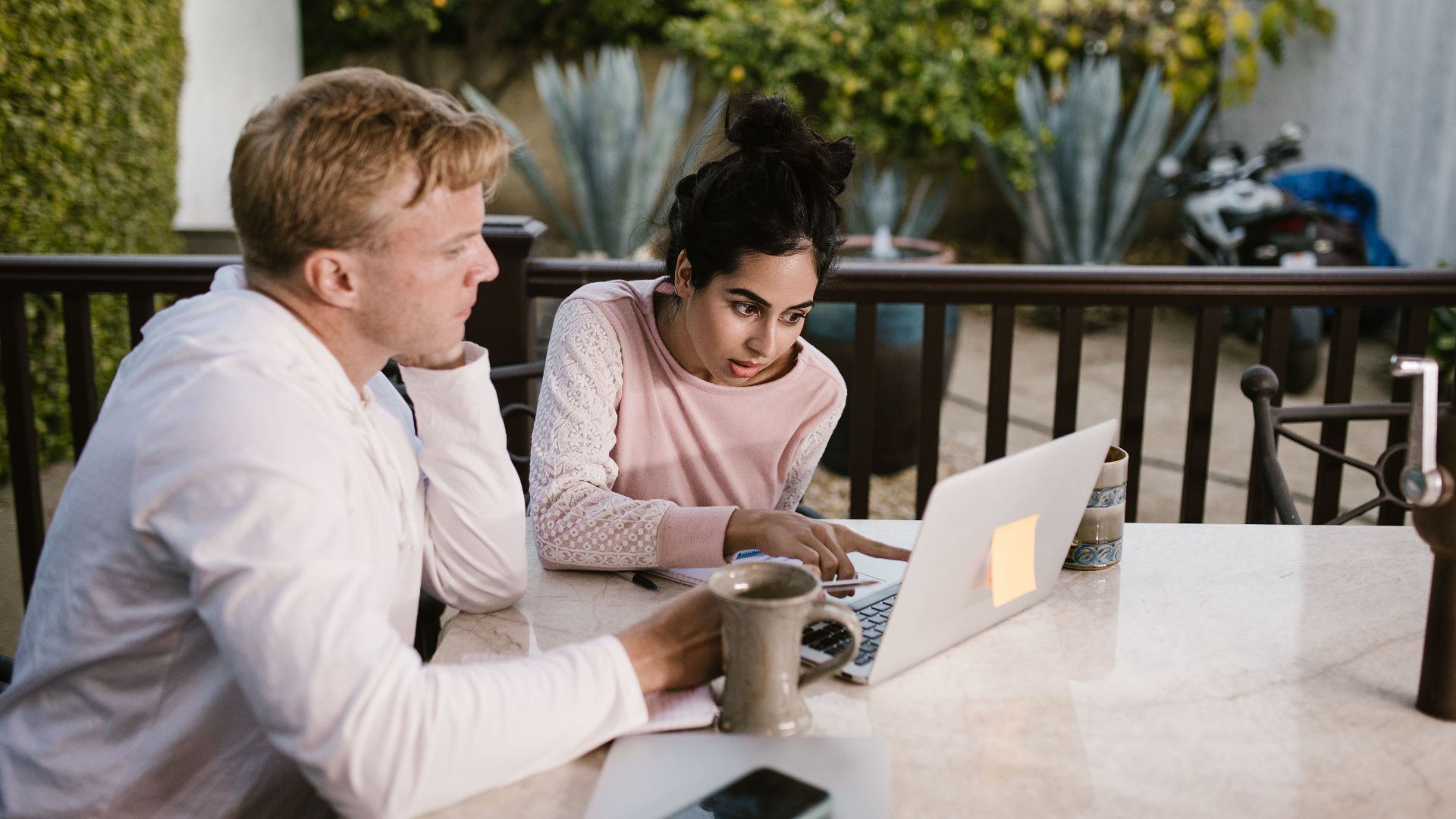 Two adults collaborating on a laptop, seated outdoors with coffee on the table.