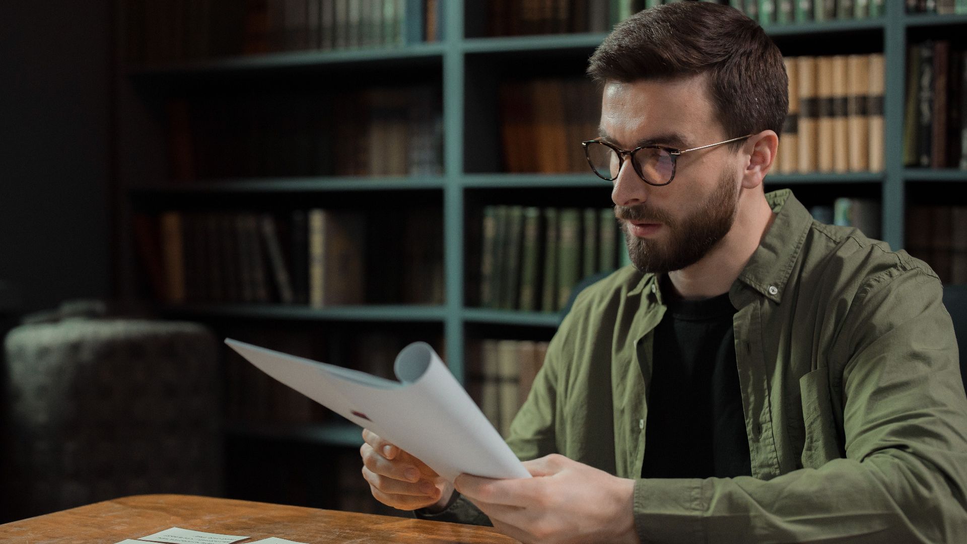 A focused man with glasses carefully reviews documents at a library desk.