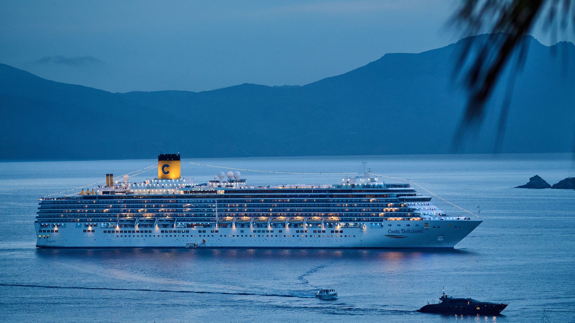 A beautifully illuminated cruise ship sails across the calm sea at dusk, framed by mountains.