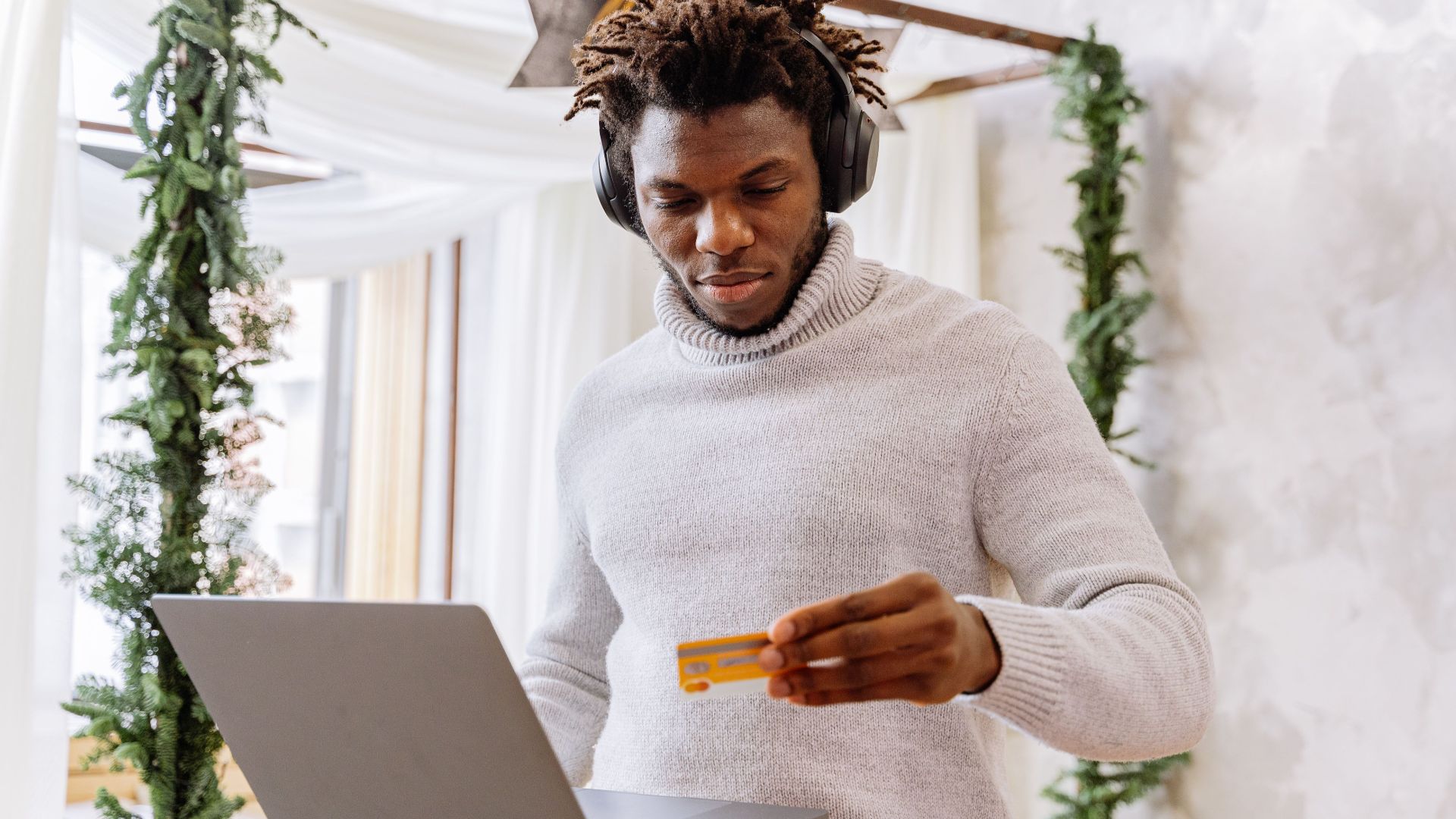 A man stands indoors with a credit card and laptop, wearing headphones and a turtleneck sweater.