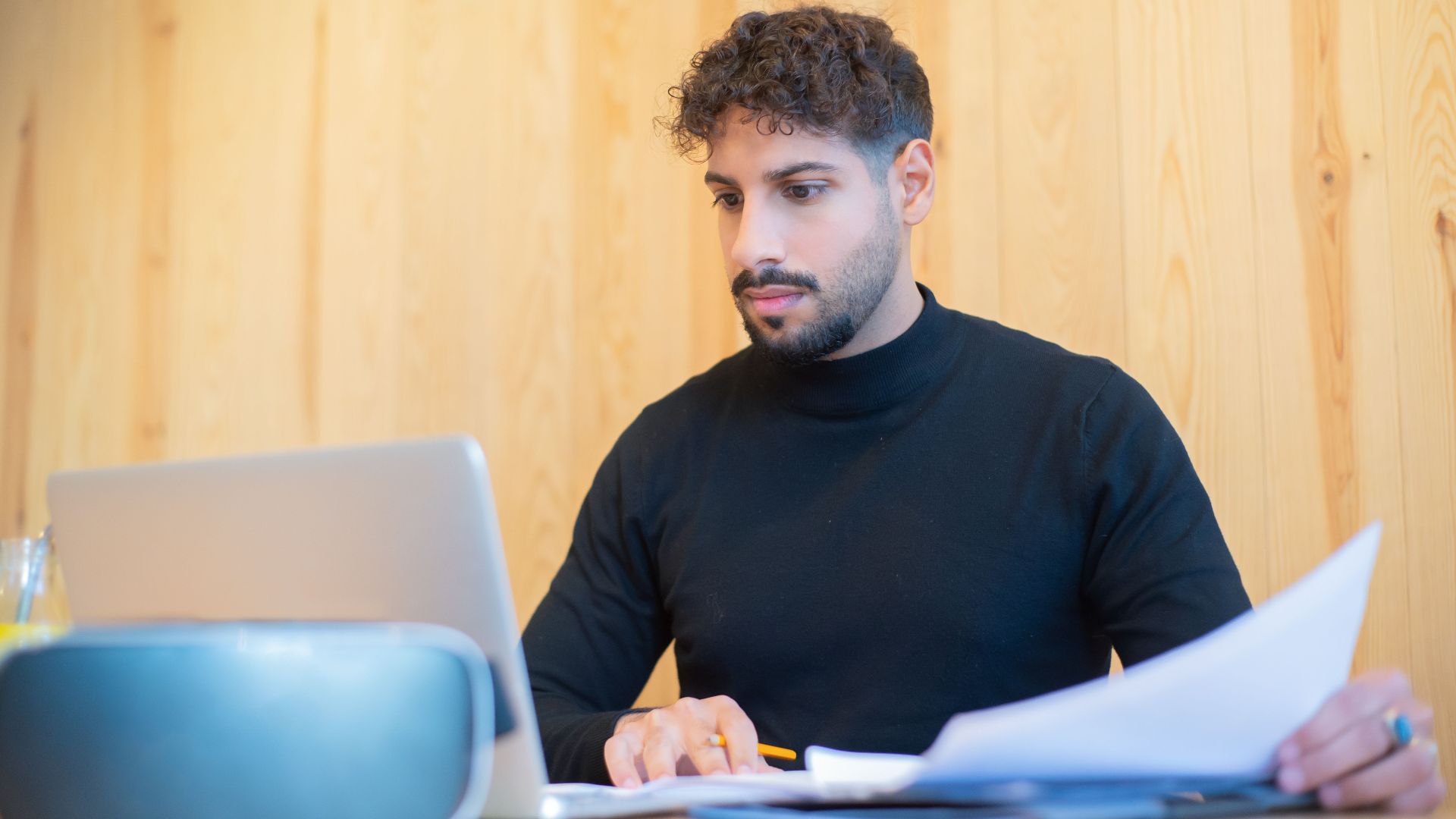 Focused businessman with beard working on a laptop, reviewing documents in a modern workspace.
