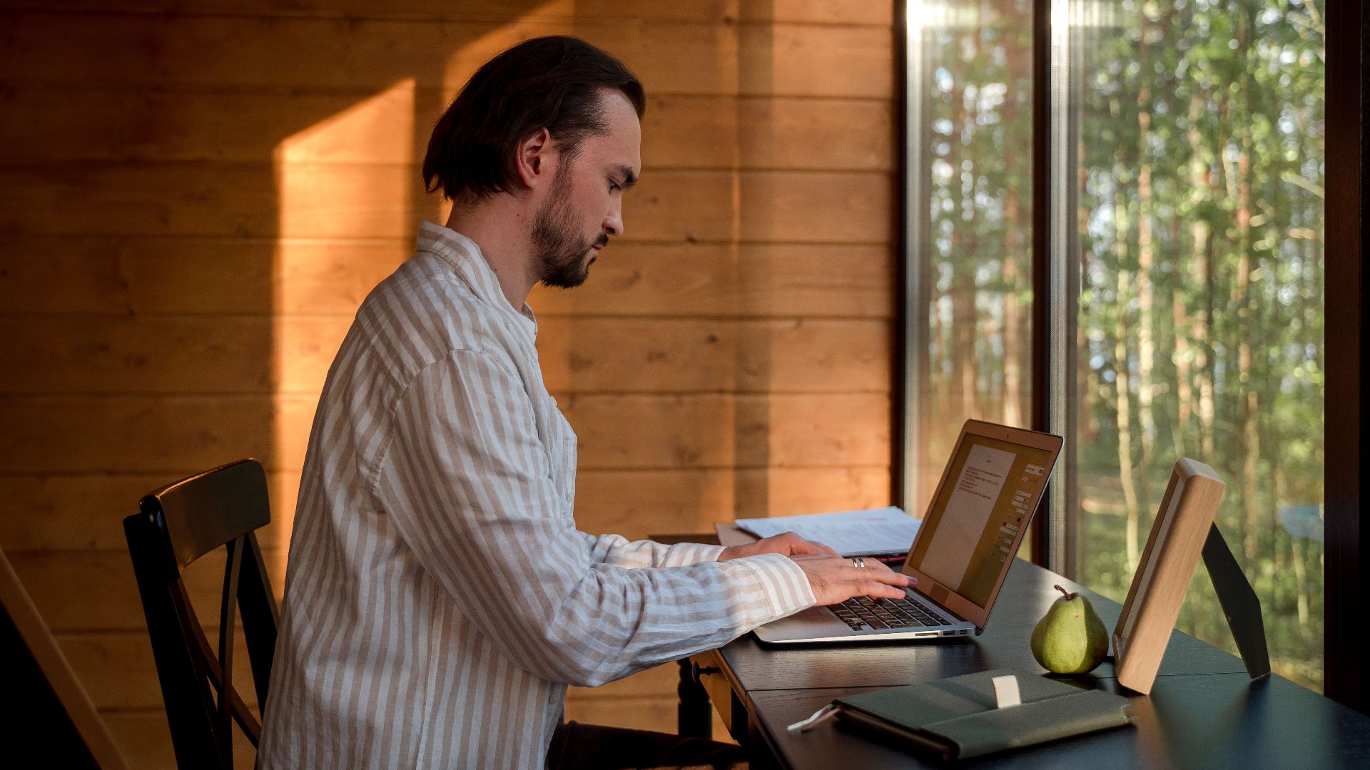 Side view of a man with facial hair typing on a laptop indoors by a window, illuminated by soft, natural light. Modern home office setup with books and a notebook on the desk.