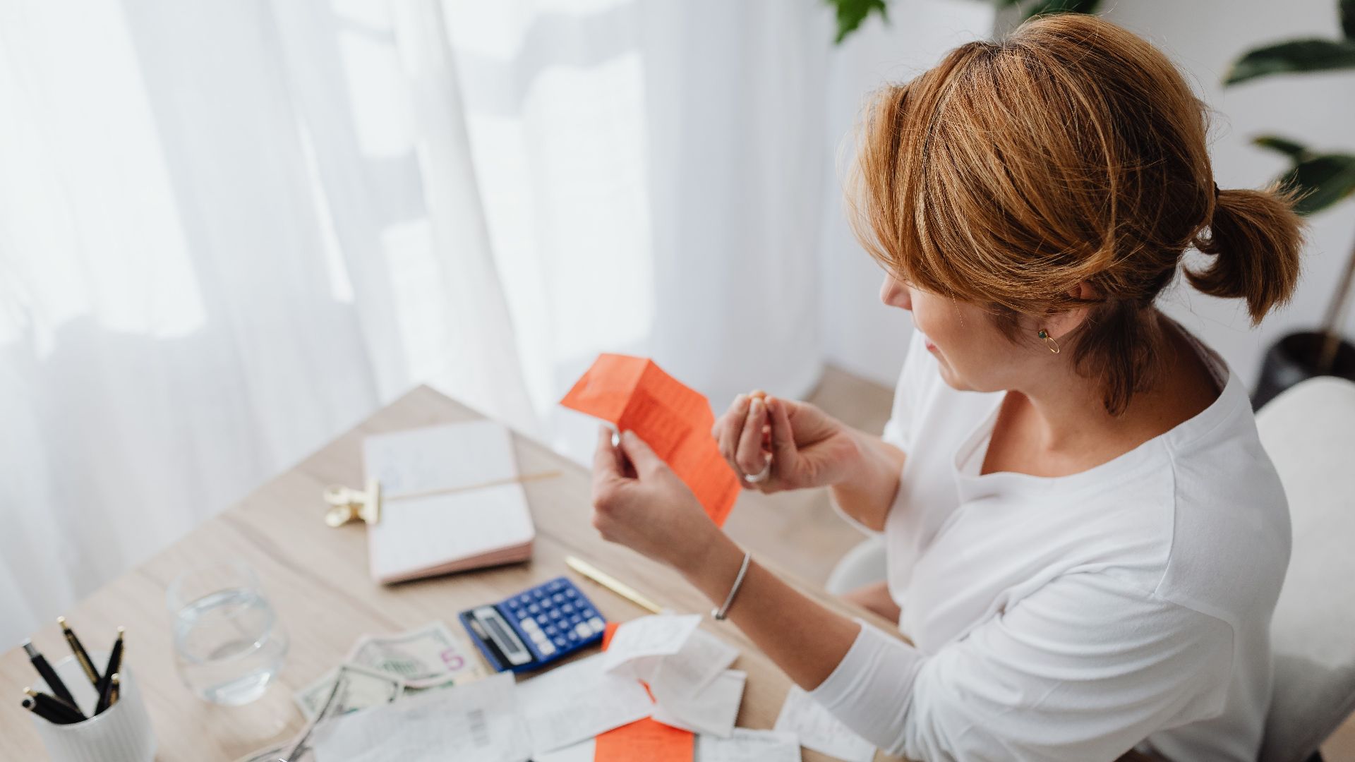 A woman organizing her finances with bills and receipts at a home office desk using a calculator and laptop.