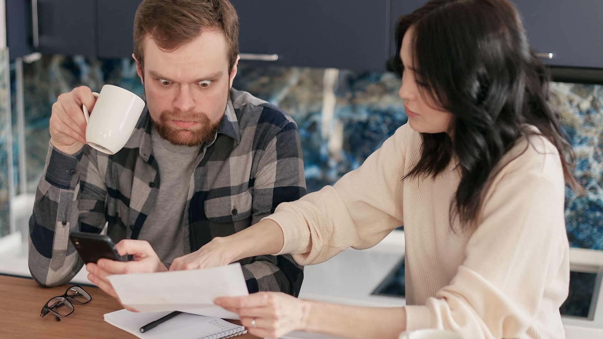 A couple reviews household bills in a kitchen, appearing concerned and focused.
