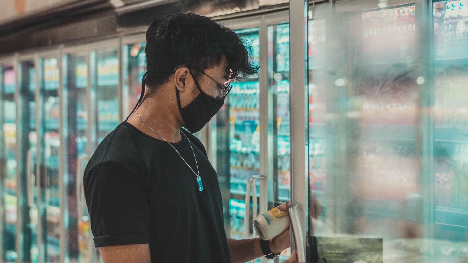 Side view concentrated young male in black shirt and face mask opening fridge door and choosing goods while doing grocery purchases in supermarket