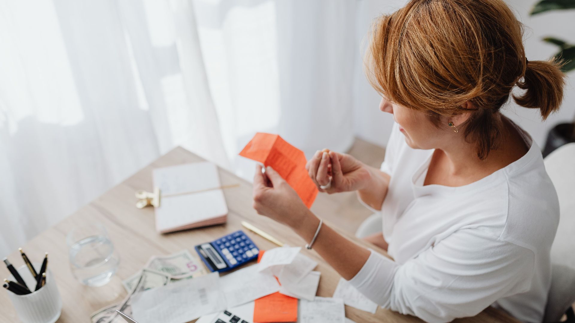 A woman organizing her finances with bills and receipts at a home office desk using a calculator and laptop.