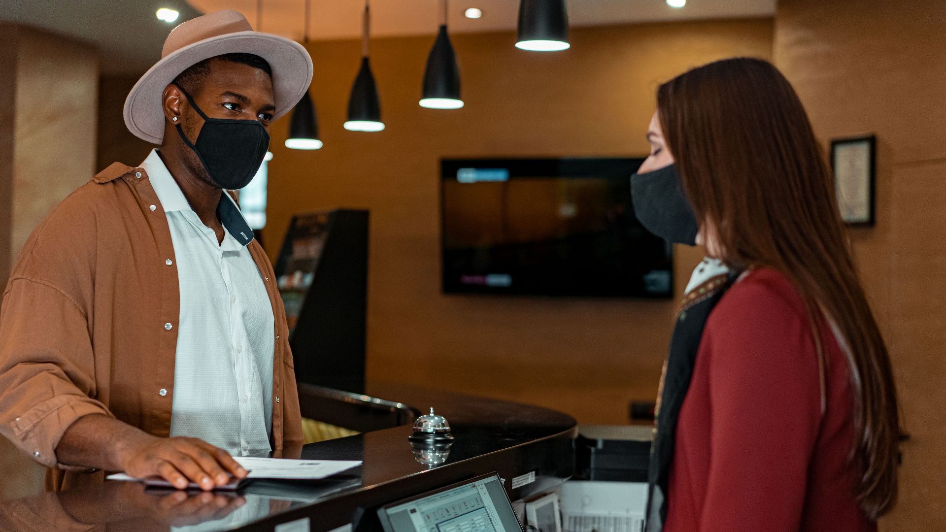A man and woman in face masks checking in at a hotel reception, emphasizing safety protocols.