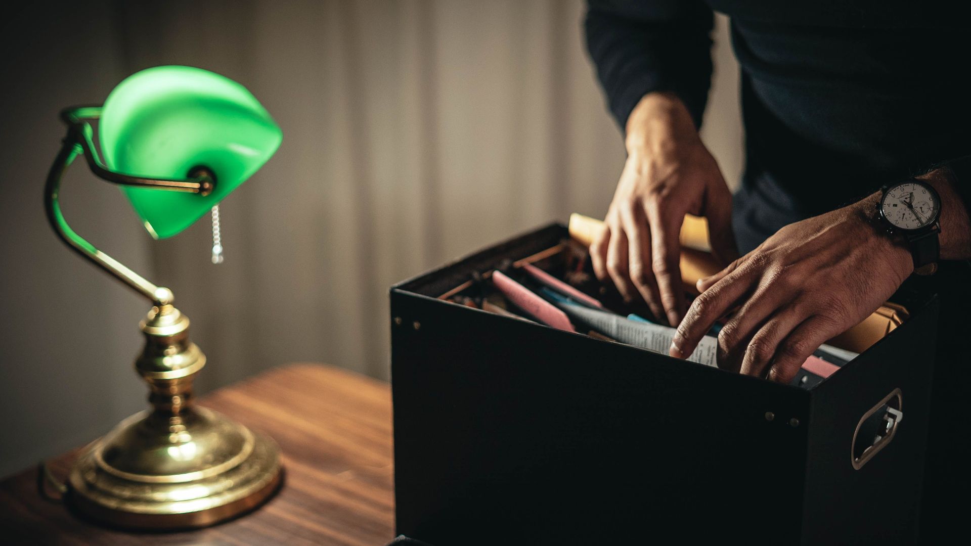 Crop anonymous male looking through files and folder in box placed on desk with green bankers lamp in home office