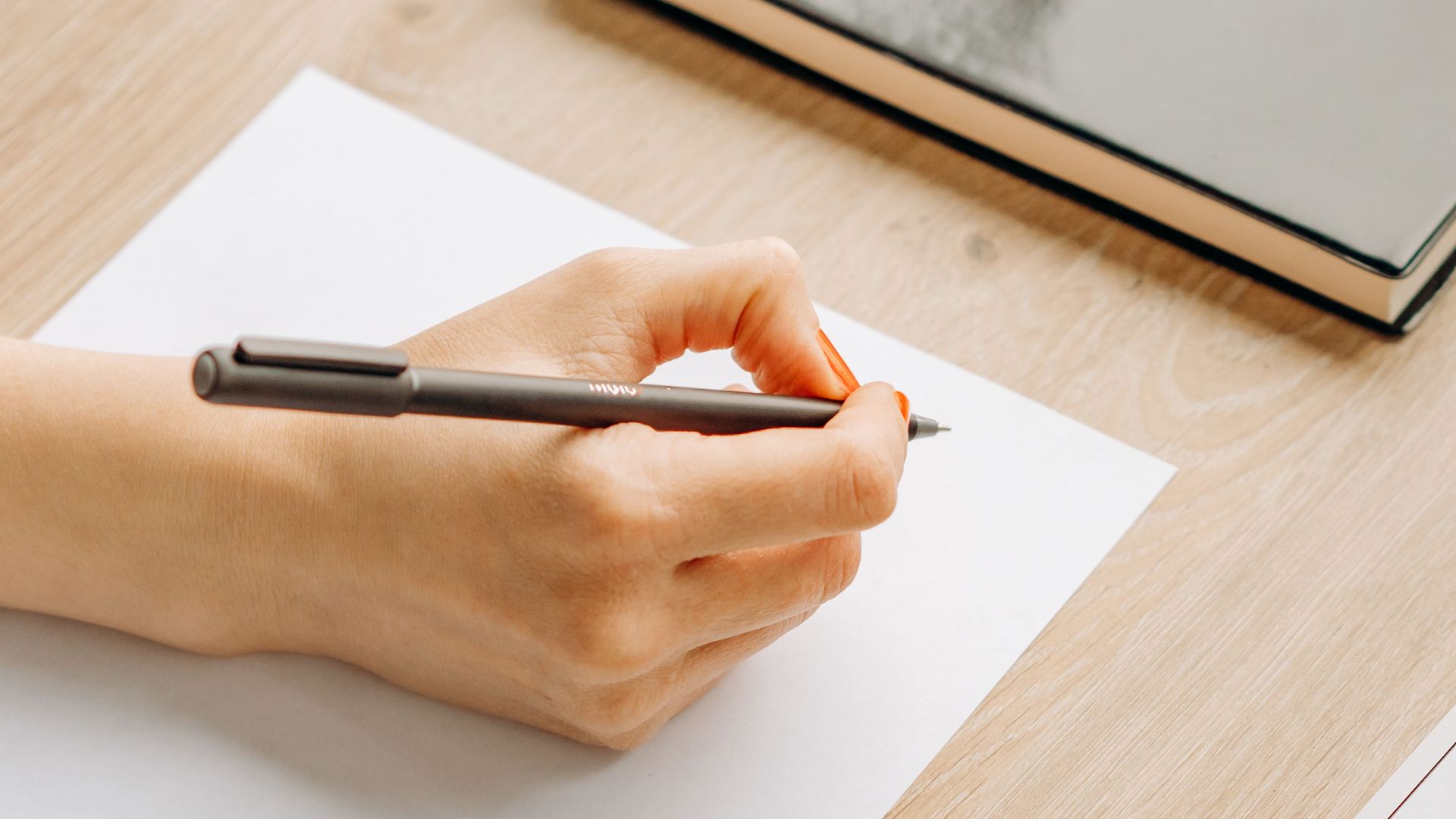 Close-up of hands writing on paper with a pen, sitting at a wooden desk indoors.