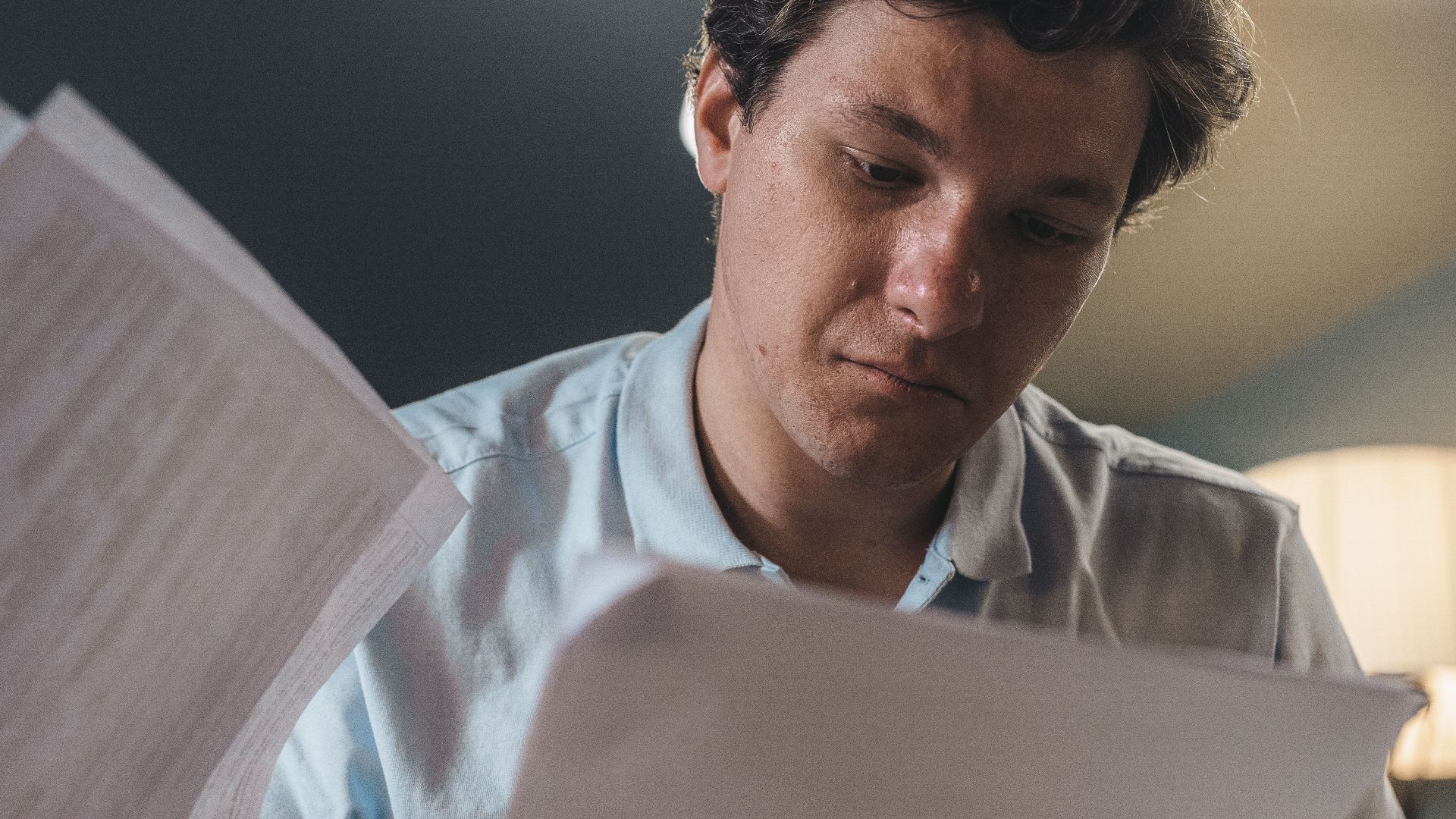 Serious man wearing a light blue shirt intently reading printed papers indoors under warm lighting.