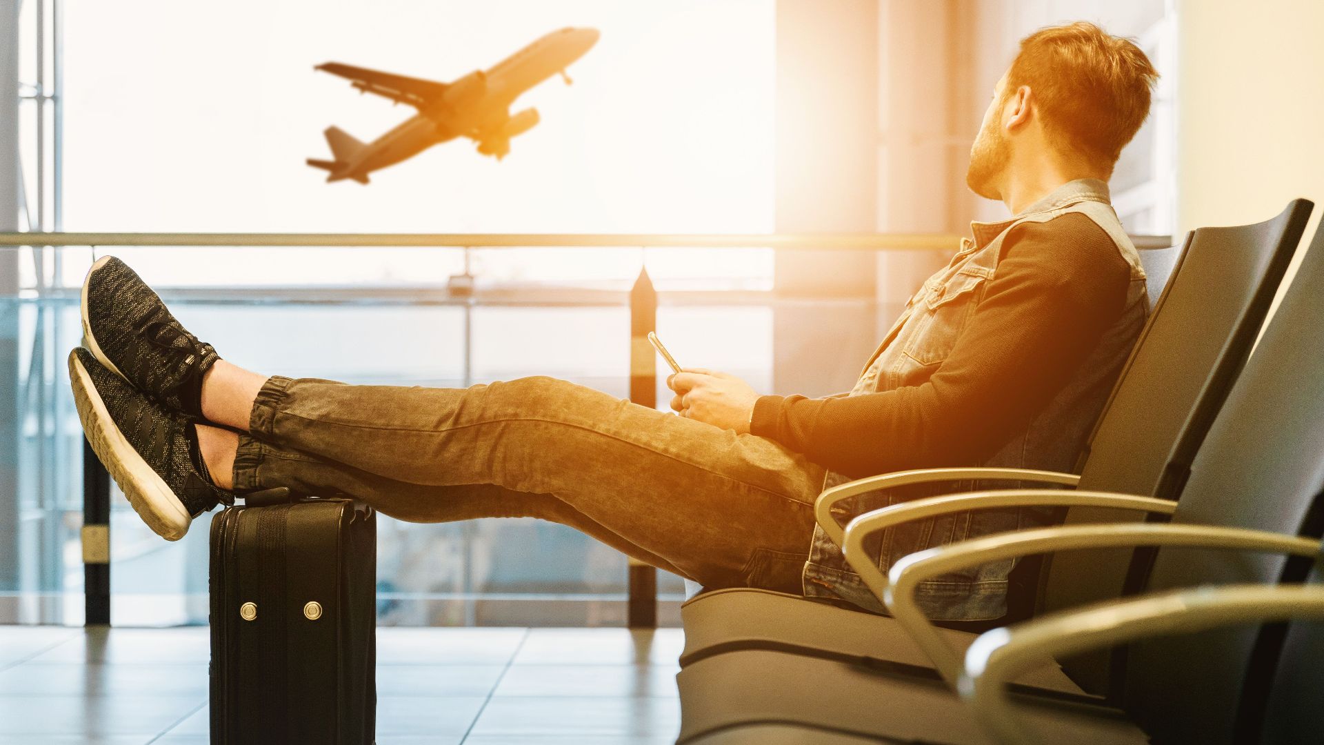 Side view full body of bearded man sitting on chair in waiting hall of airport and looking at flying aircraft behind window