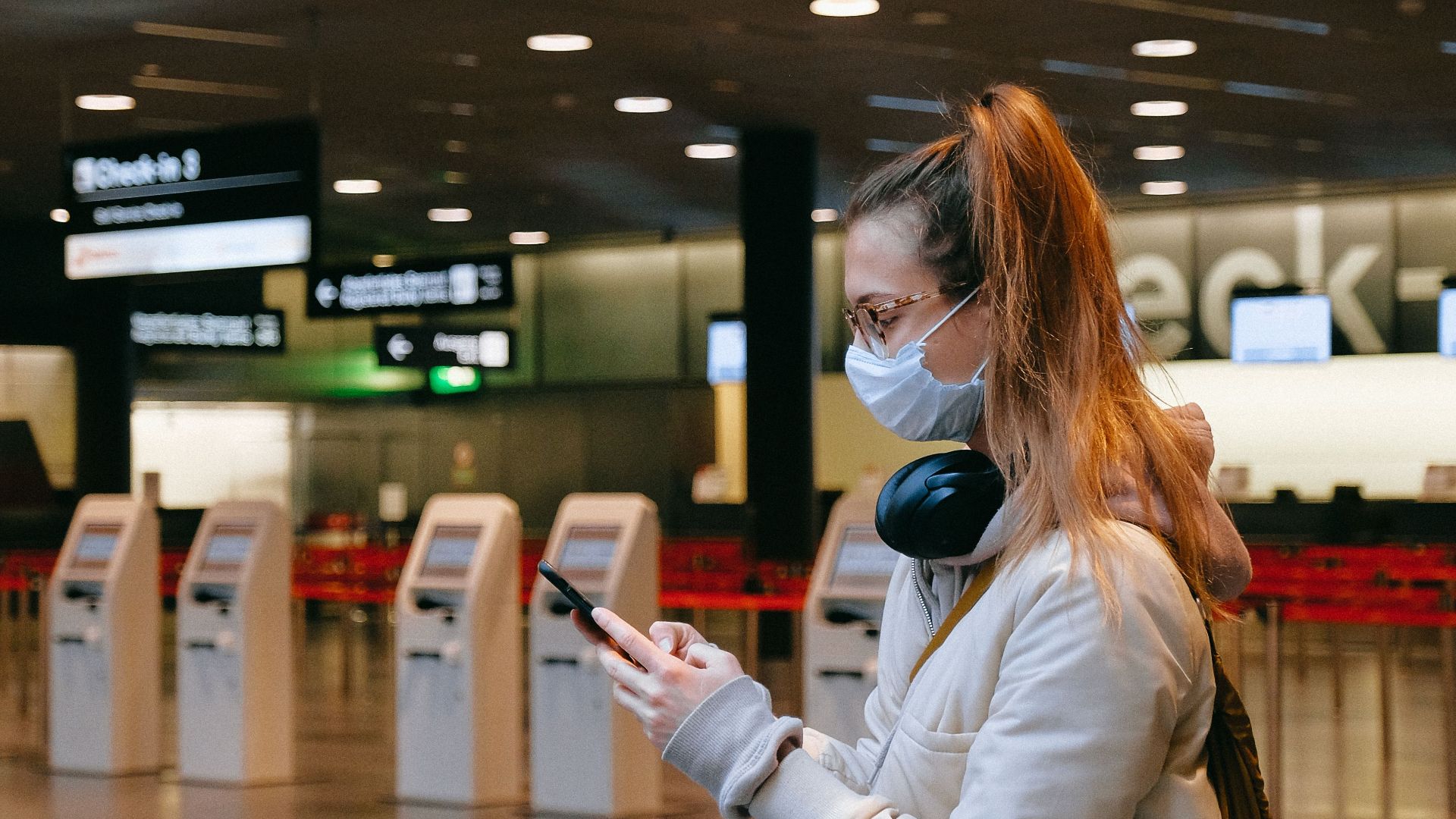 Woman wearing a mask, texting in airport terminal with signage in background.