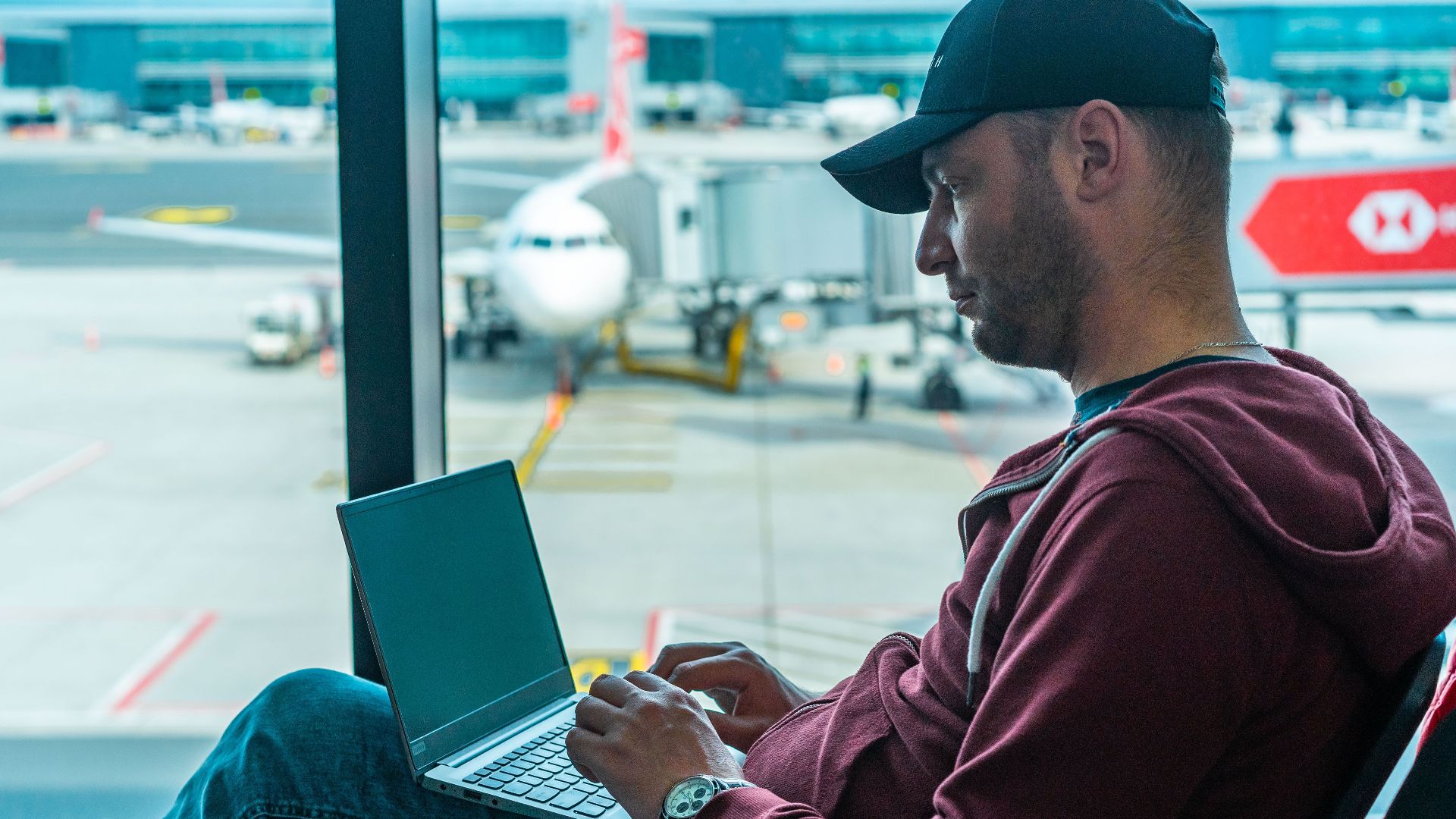 A man working on his laptop in an airport terminal, with a plane visible outside.
