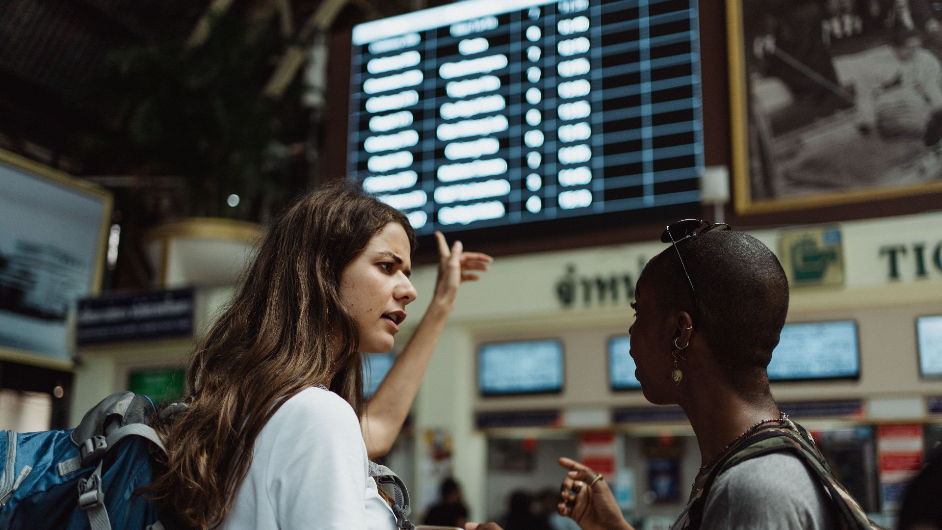 Two travelers with backpacks discuss in front of an airport timetable. Adventure awaits.