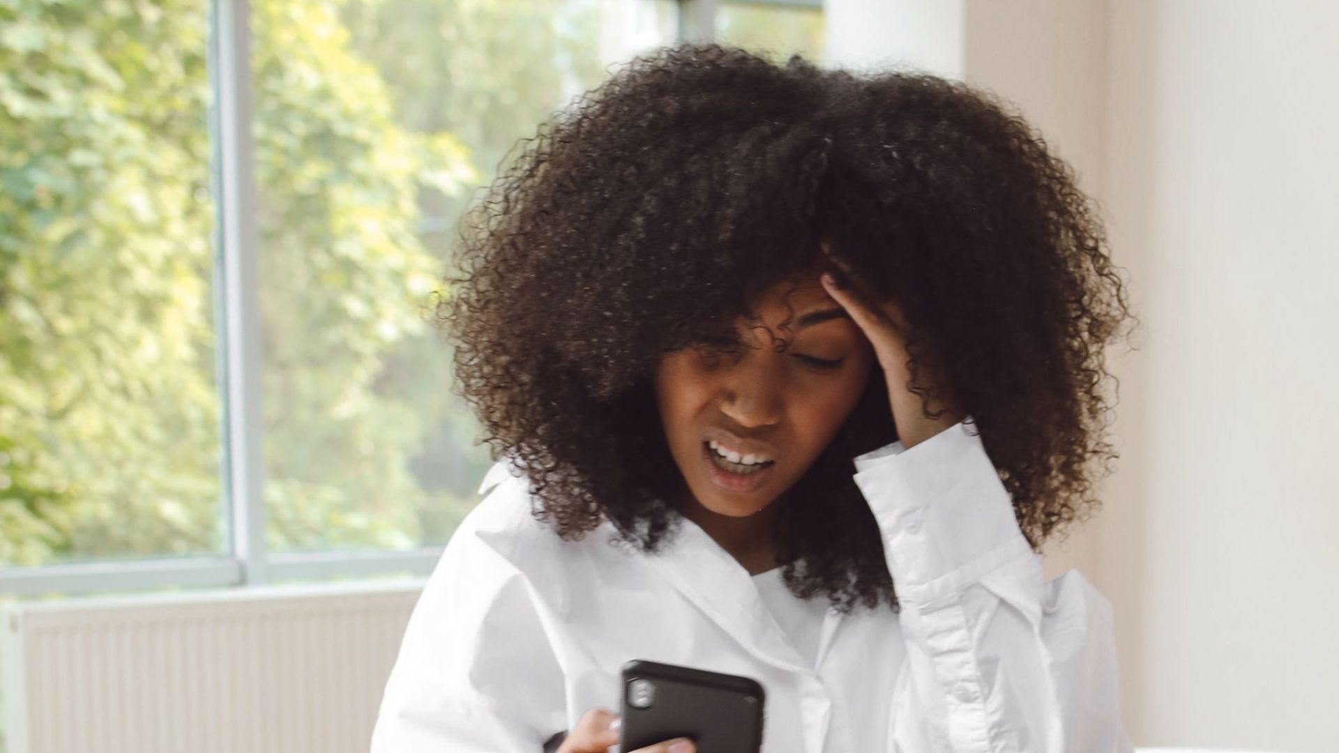 African American woman with curly hair using smartphone indoors, showing stress.