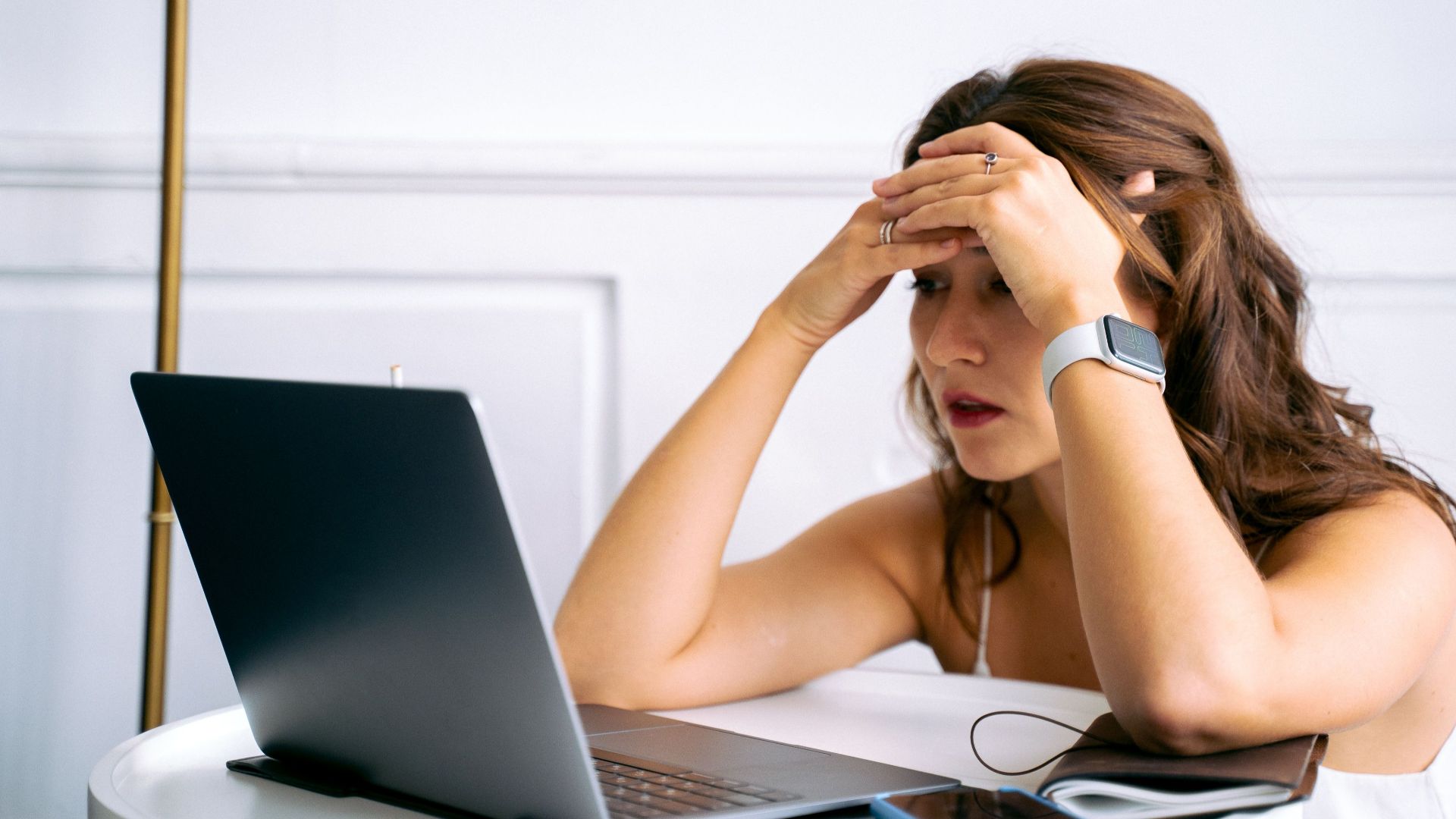 Woman feeling stressed while working on a laptop, using smartphone and smartwatch at a desk.