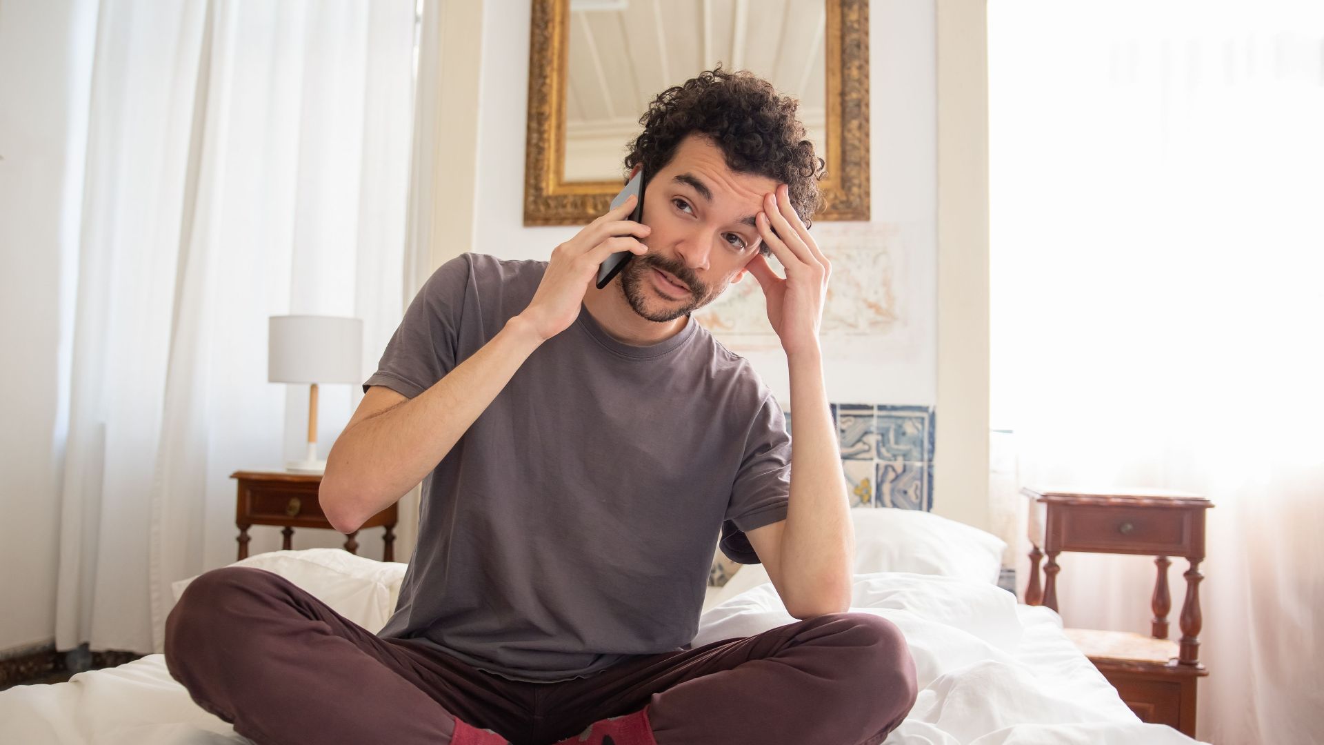 A man in casual attire sits on a bed talking on the phone with a thoughtful expression.