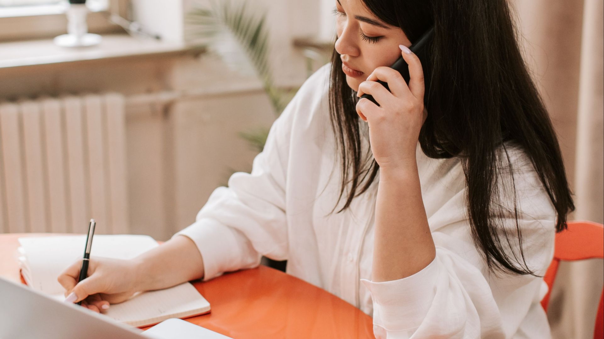 Woman multitasking in a home office, using phone and laptop at a table.