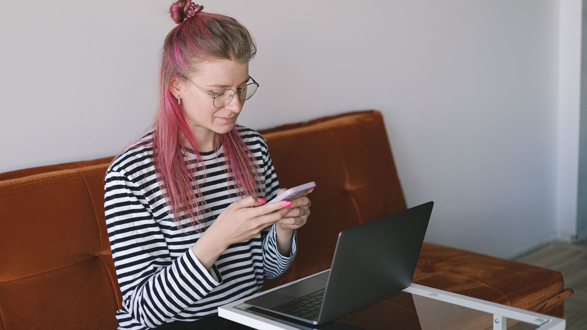 Woman with pink hair using a smartphone and laptop on a couch. Focus on technology and casual lifestyle.