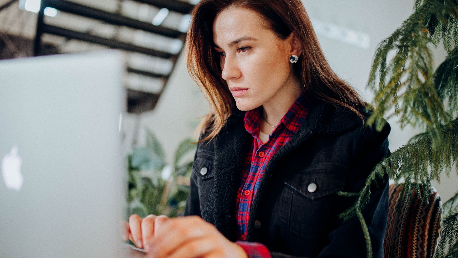 Young woman in plaid shirt intensely working on laptop in a modern environment with greenery.