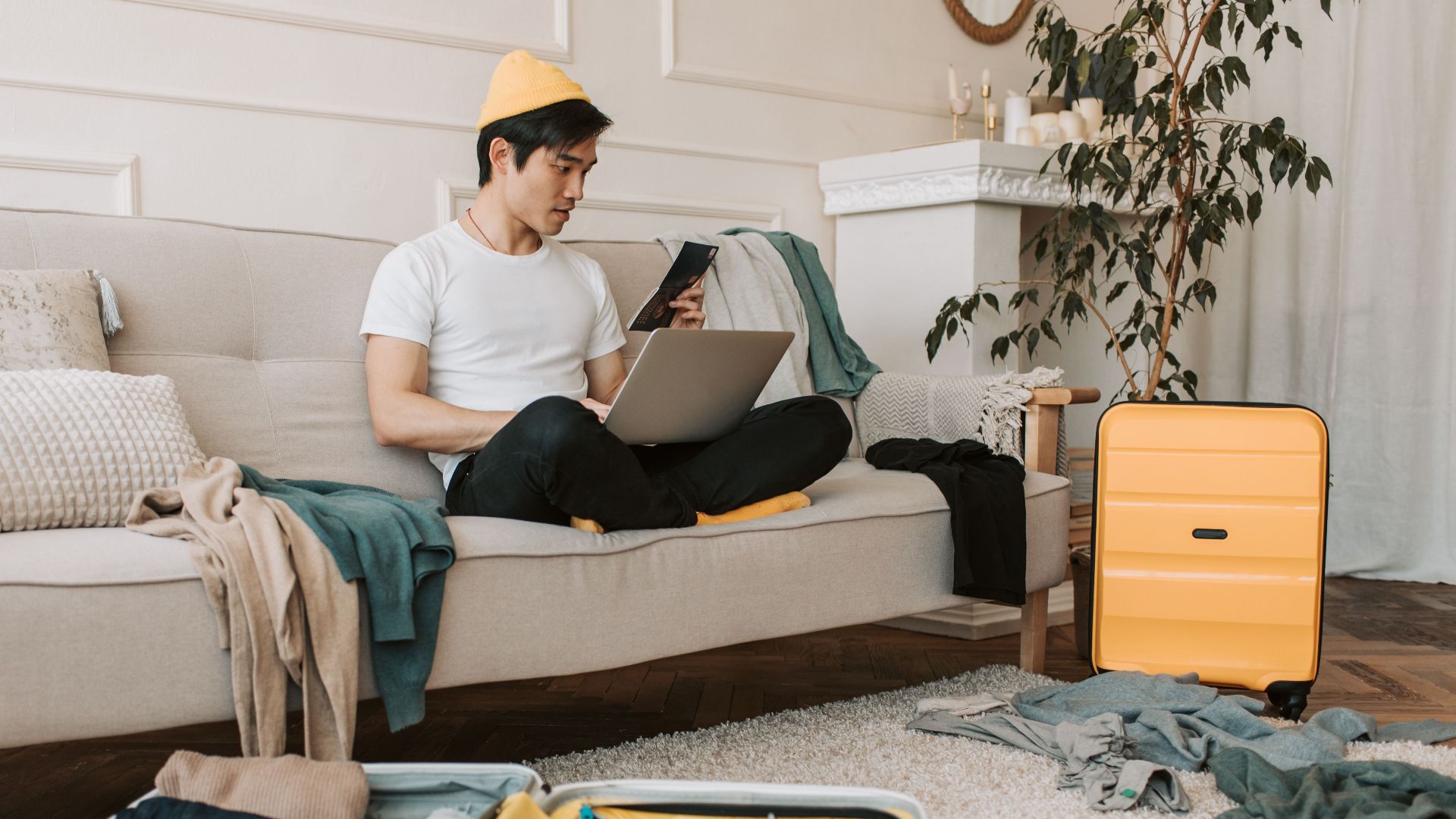 Adult male traveler planning trip at home with laptop and smartphone, surrounded by luggage.