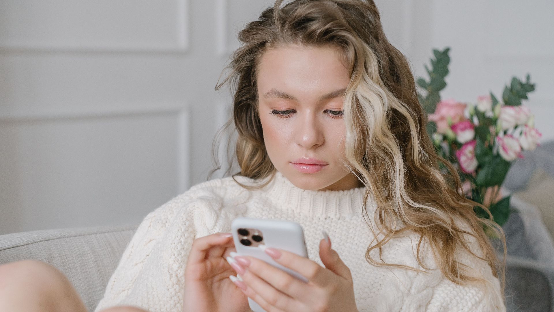 Young woman in a sweater sitting on a sofa, focused on texting with her smartphone indoors.