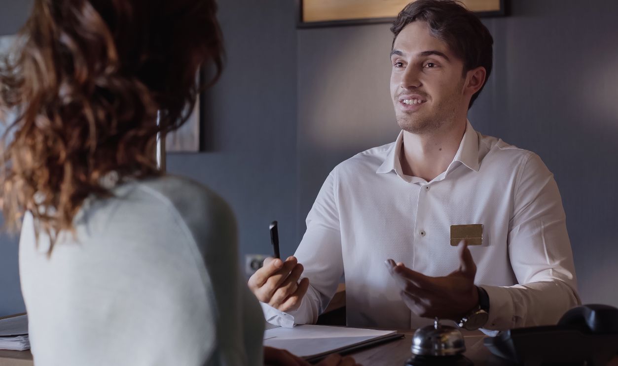 Shutterstock-2061881507, smiling hotel receptionist talking with blurred woman at hotel counter