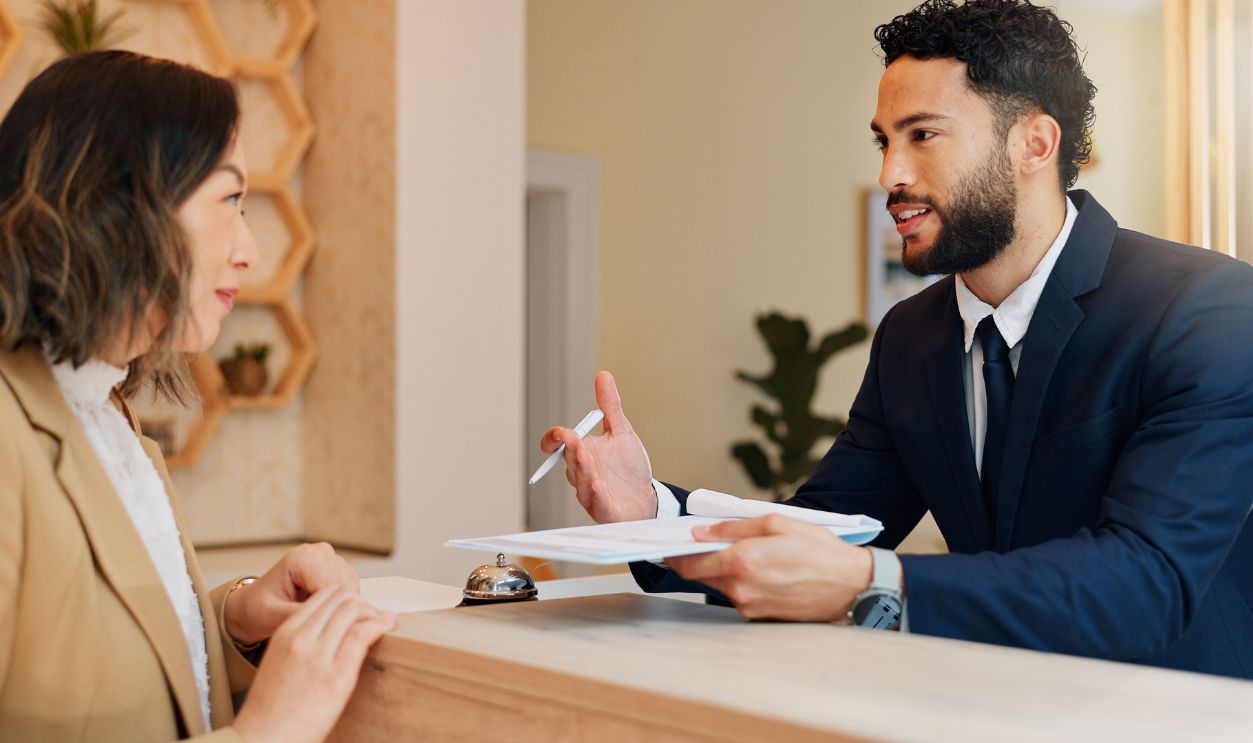 Shutterstock-2573105107, Hotel reception, info and woman at desk with man, paperwork and friendly service for hospitality. Check in, registration and receptionist with client at counter for accommodation, booking or travel