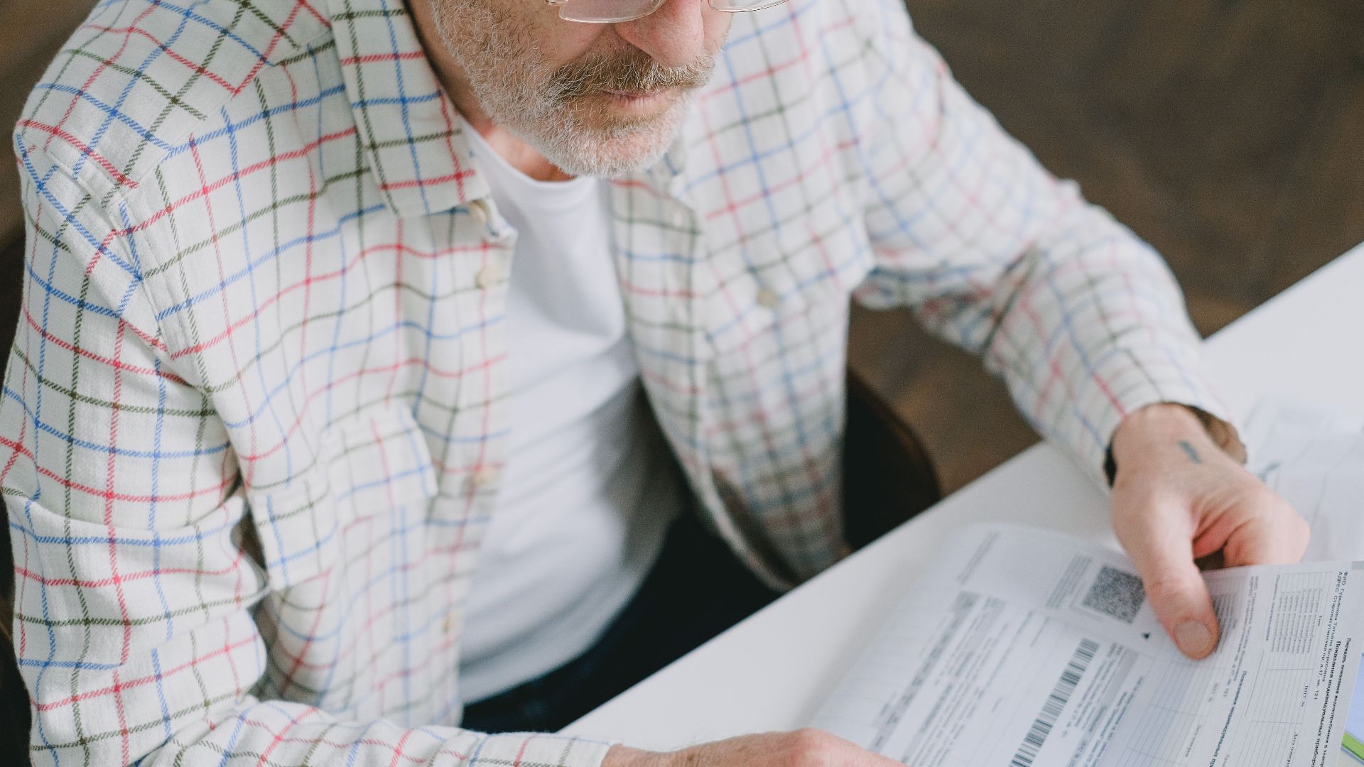 Elderly man in glasses carefully examines documents at home. Thoughtful and focused.