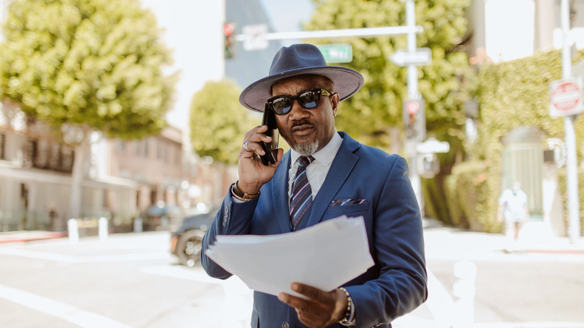 A confident businessman in a blue suit and hat talks on the phone while holding documents outdoors.