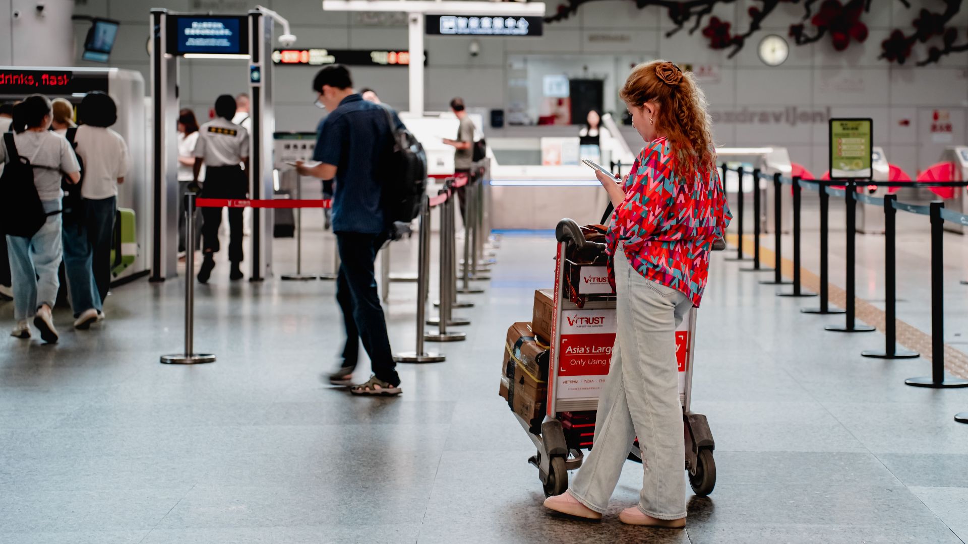 A traveler checks her phone while waiting with a luggage cart at a busy airport terminal in Guangzhou.