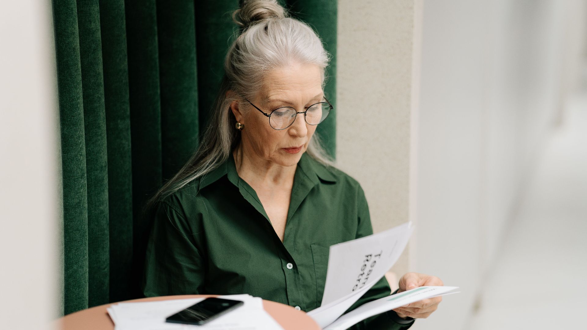 Elderly woman with gray hair reading papers at a small table indoors, focused and thoughtful.