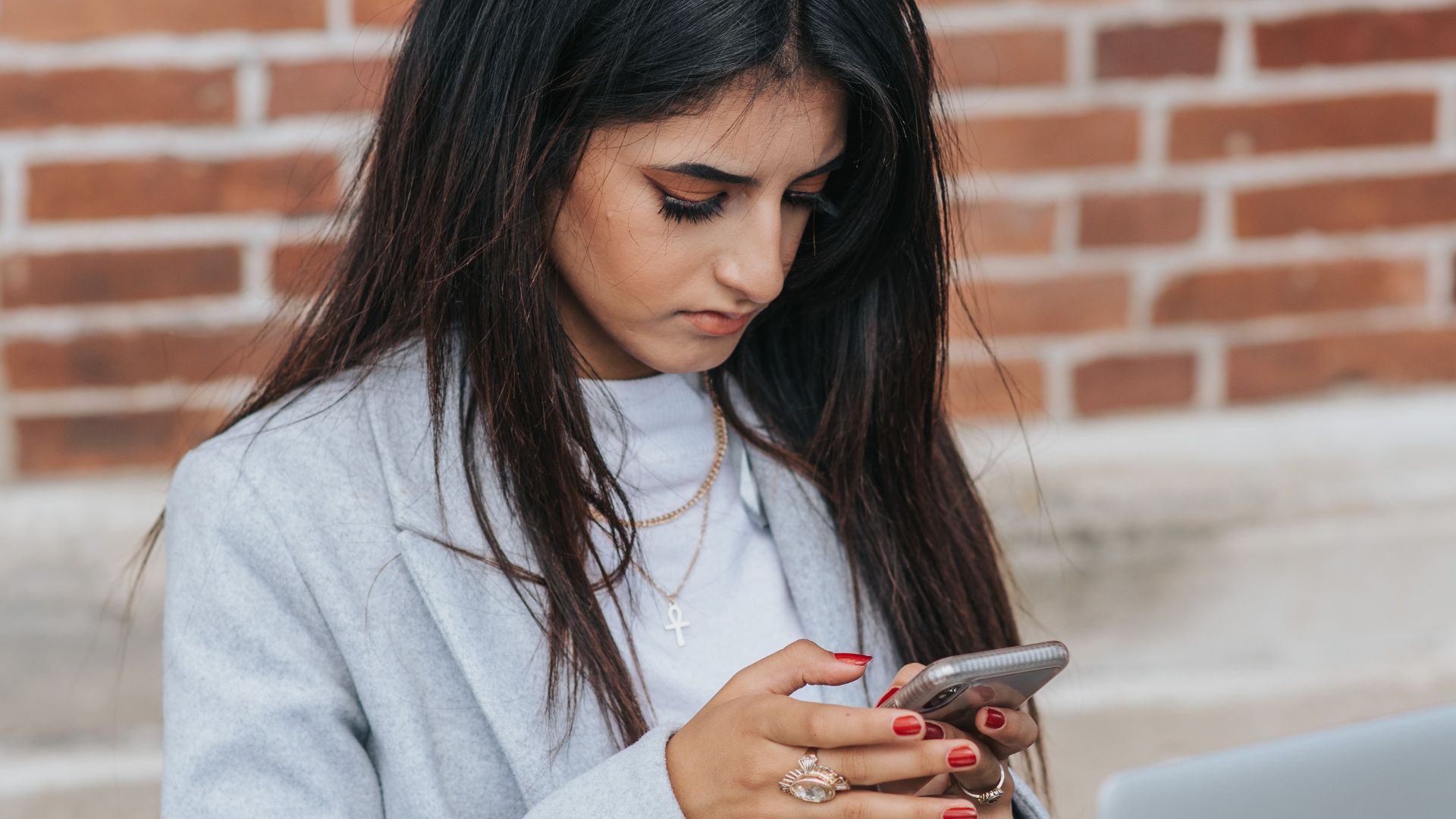 Young attentive ethnic female freelancer text messaging on cellphone near portable computer on table in town