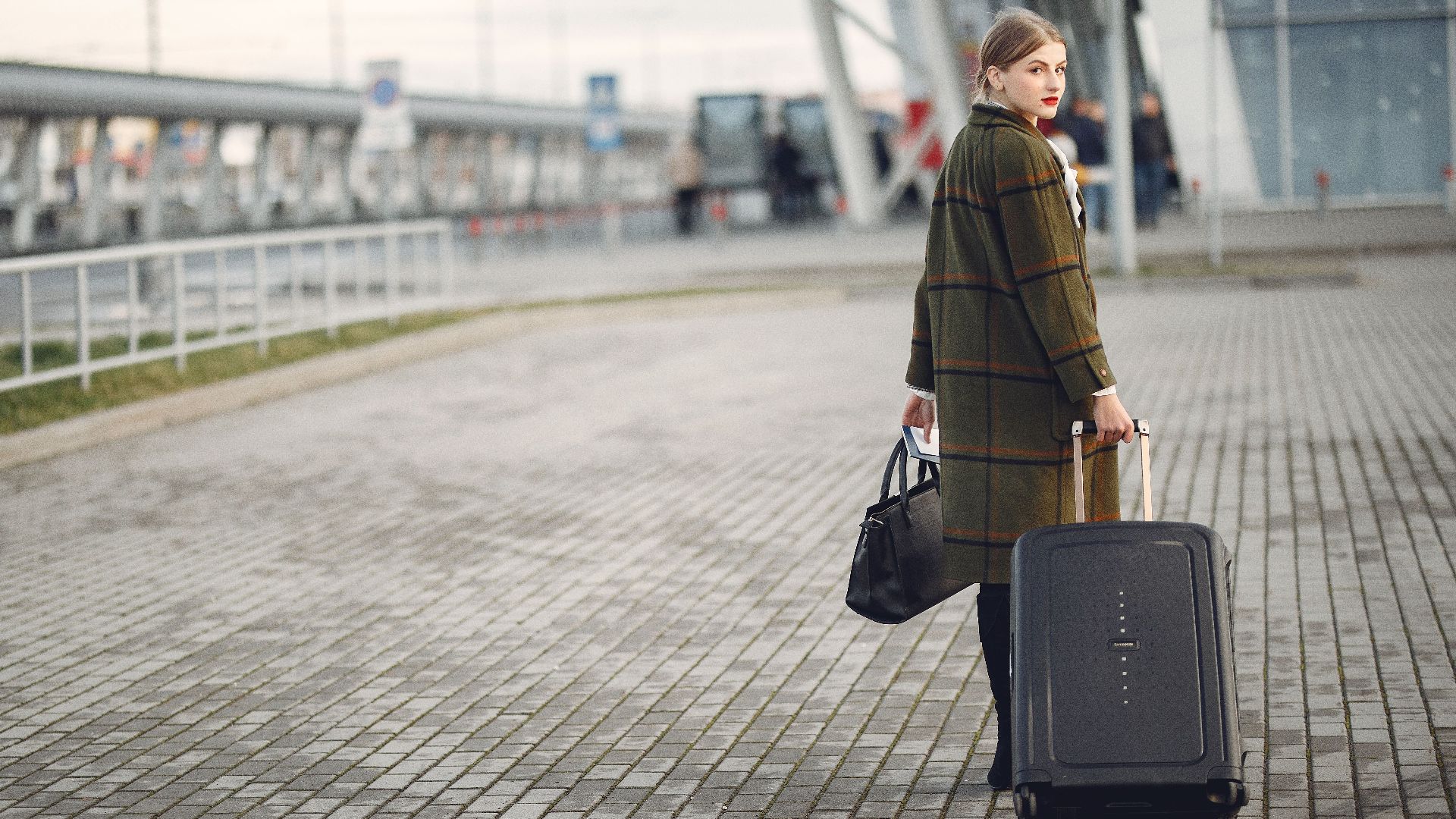 Full length of stylish female traveler in warm coat walking with suitcase and bag looking at camera after arrival against airport terminal