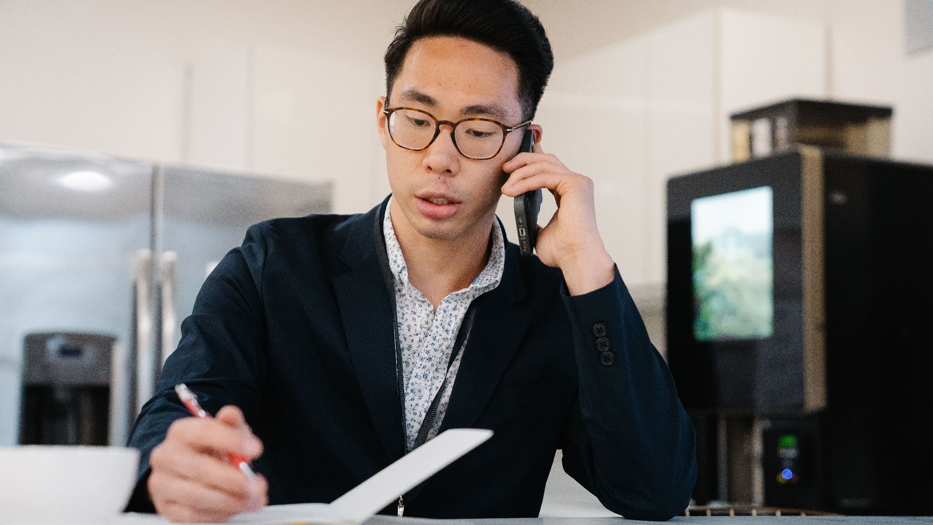 Asian man in a suit jacket on a phone call, taking notes in an office.