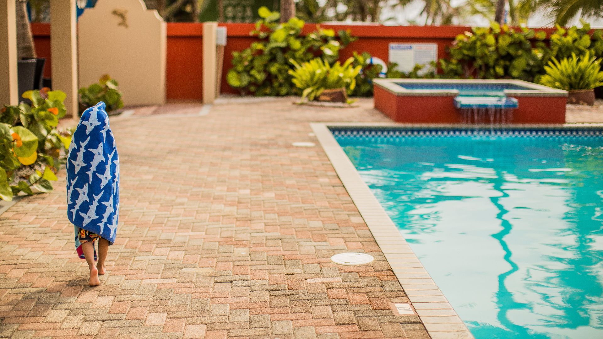 A child wrapped in a blue towel walks poolside at a tropical resort.