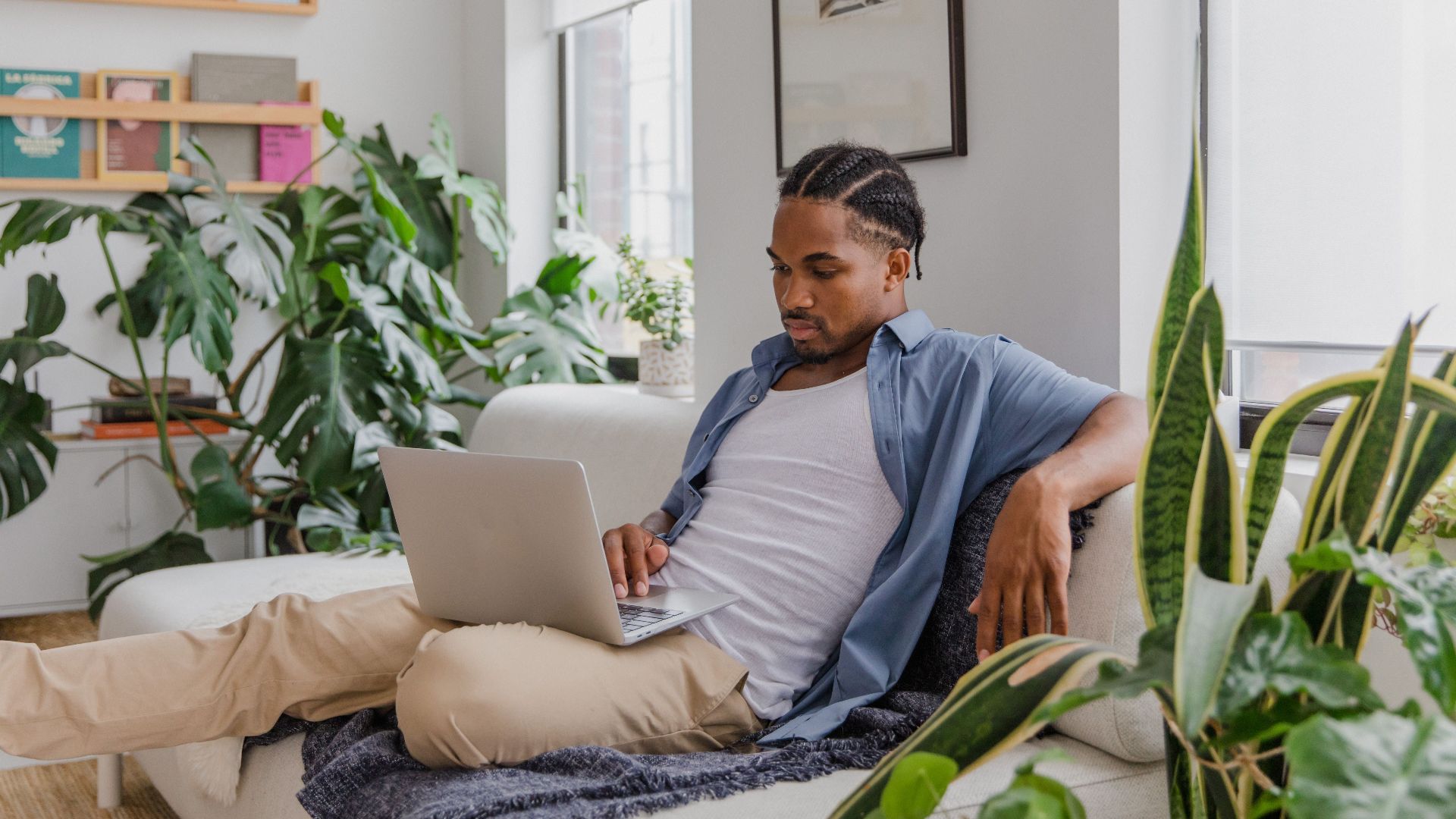 Casual man working from home office, surrounded by plants, on a comfortable sofa with a laptop.