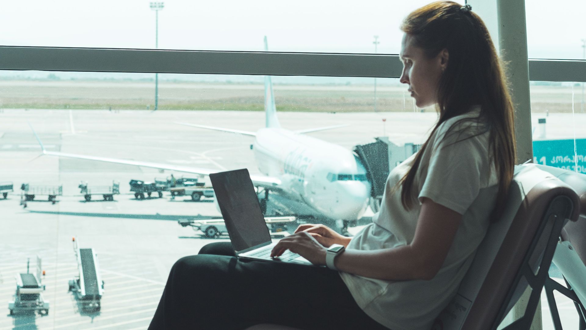 Side view of woman using laptop at airport, airplane visible outside through large glass windows.