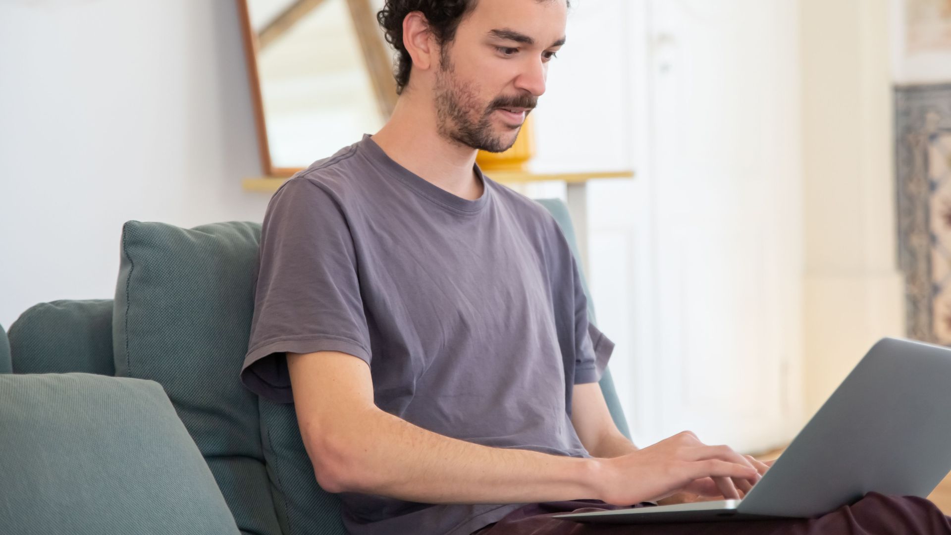 A man with curly hair typing on a laptop while sitting on a comfortable couch at home.