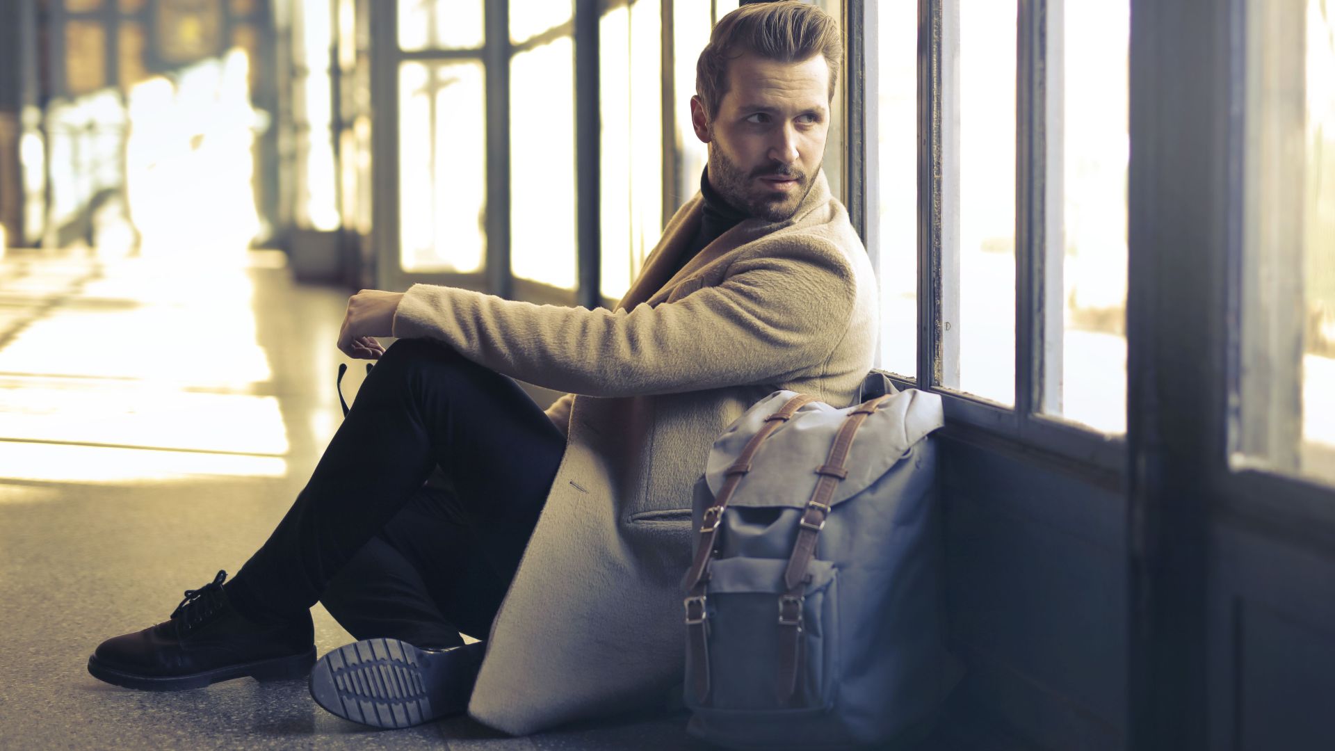 Young man in a stylish coat sits indoors with a backpack, gazing thoughtfully by large windows.