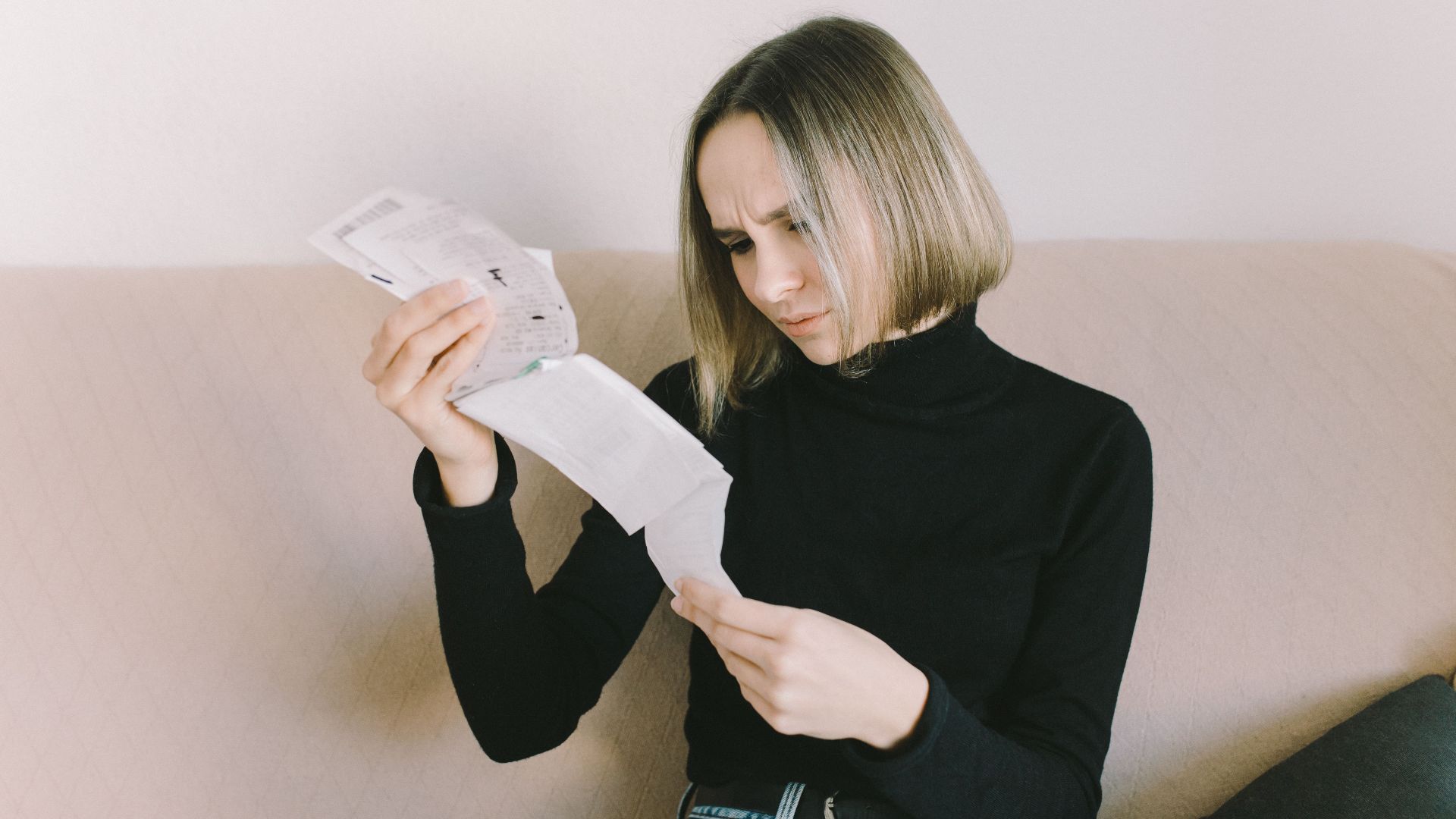 Concerned woman in black sweater examining bills on beige sofa indoors.