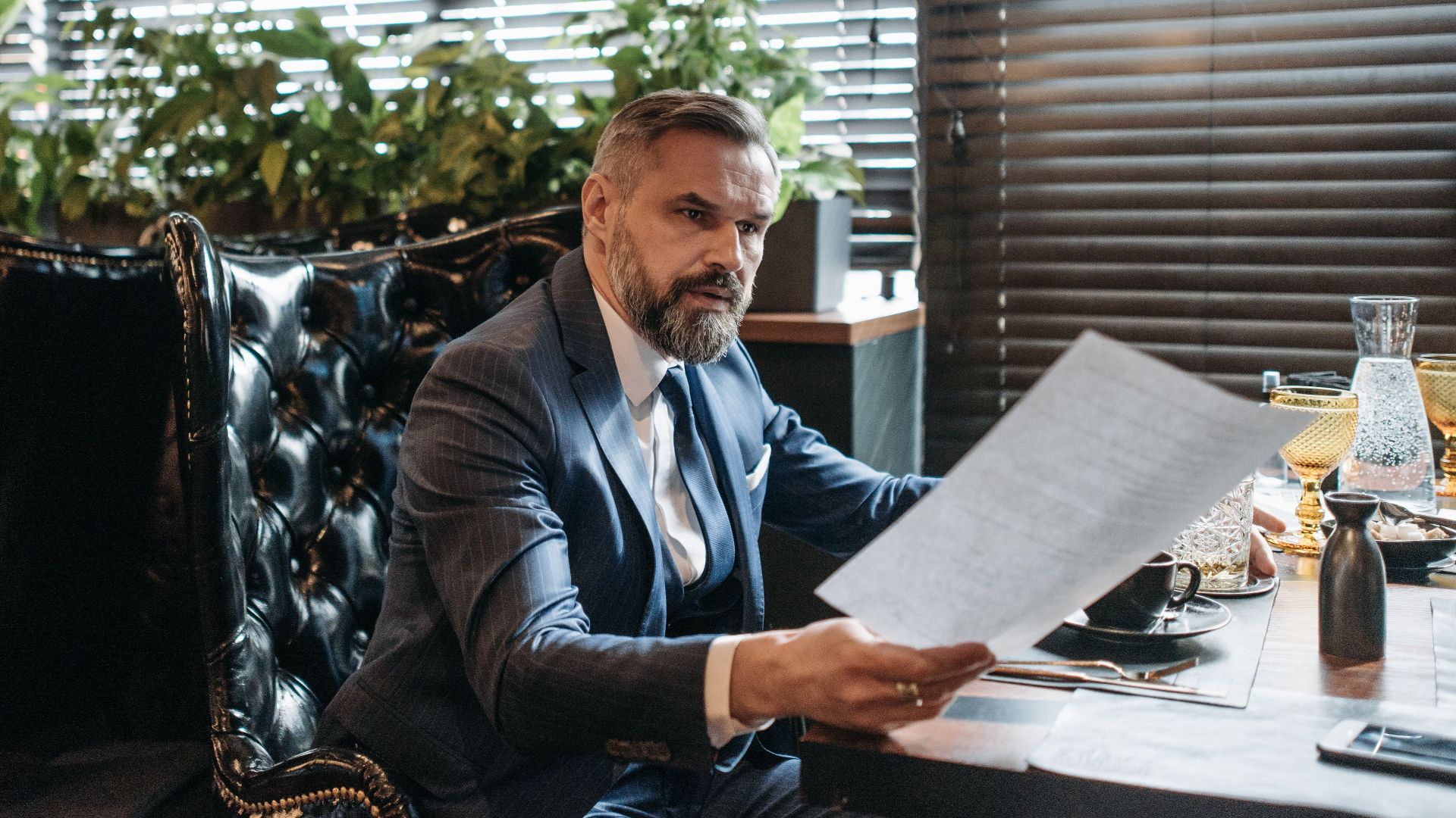 Elderly businessman in a suit analyzing paperwork in a modern office with plants.