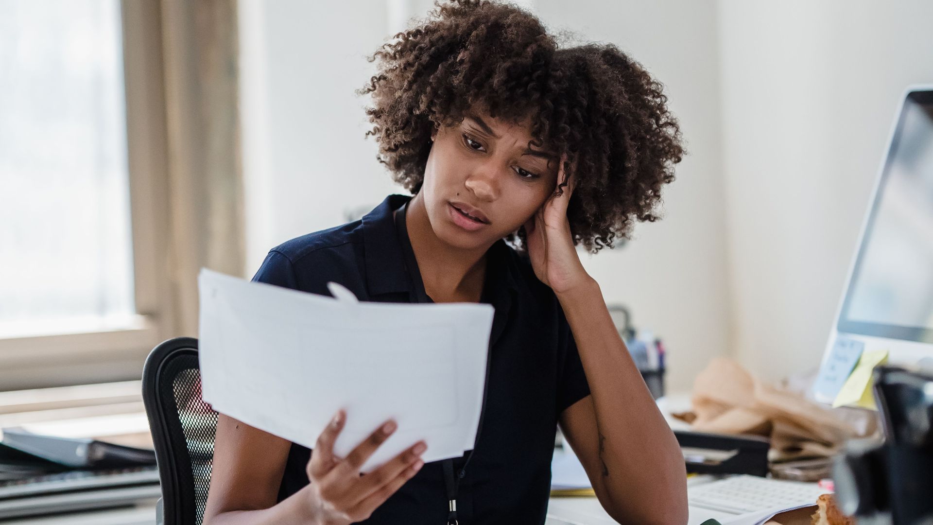 A professional woman with curly hair reviews documents at her office desk, deep in thought.