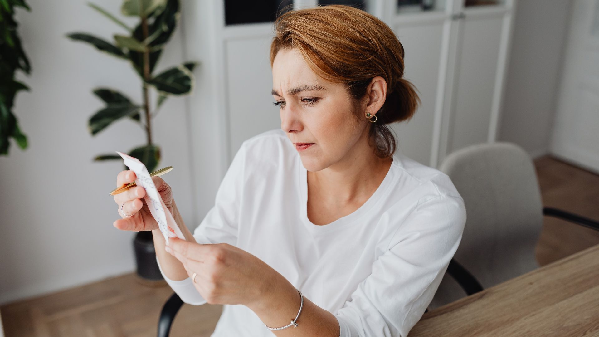 A concerned adult woman examines a receipt while sitting at her office desk, indicating financial scrutiny.