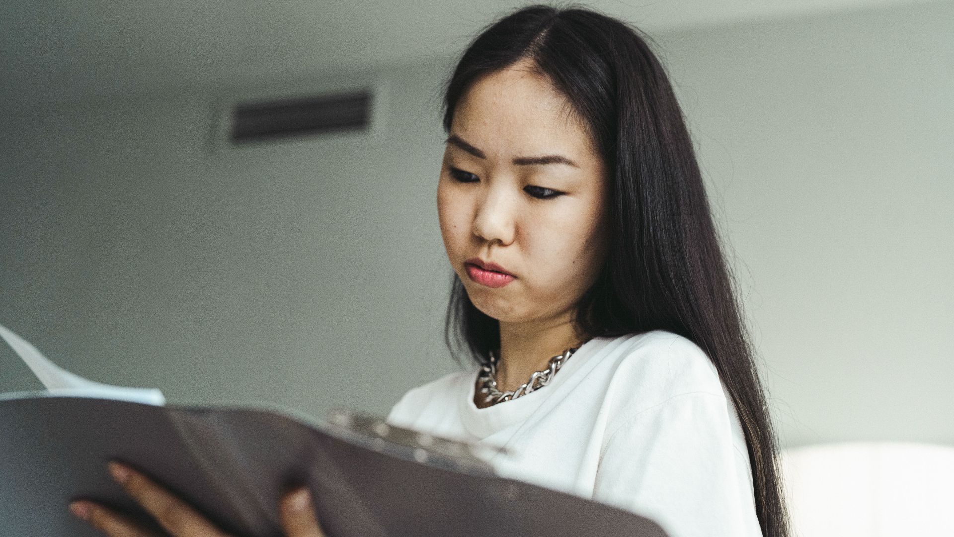 Asian woman in casual attire examining paperwork with concentration indoors.