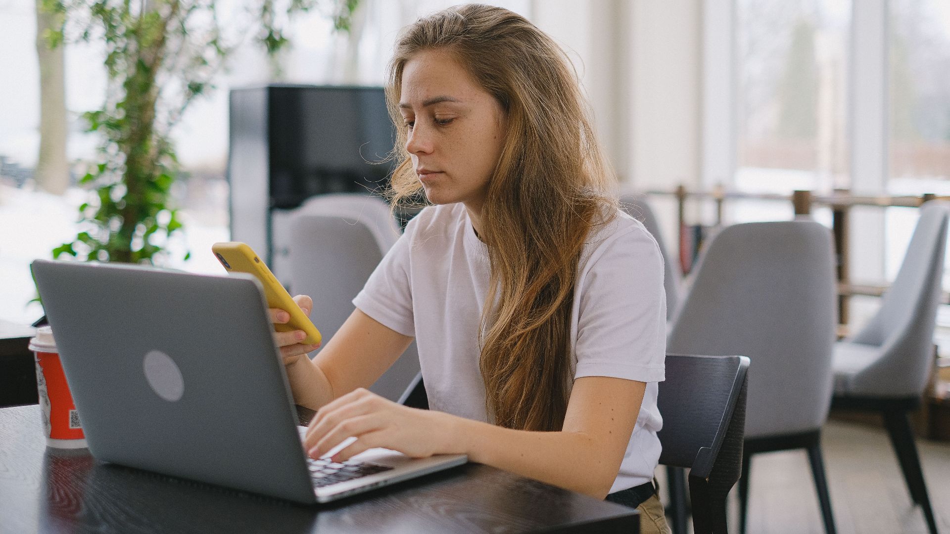 Focused woman using a laptop and smartphone at a cafe, showcasing modern multitasking.