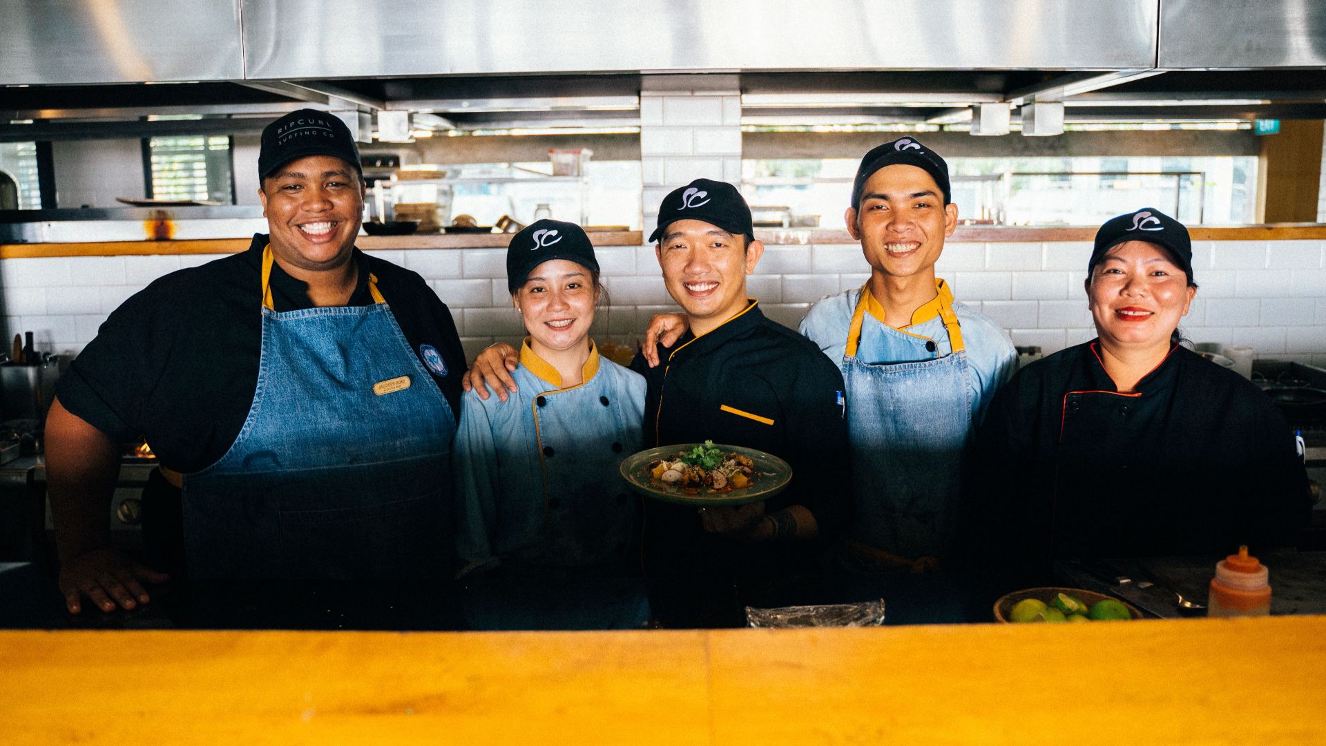 A group of diverse chefs smiling and posing together in a modern restaurant kitchen.