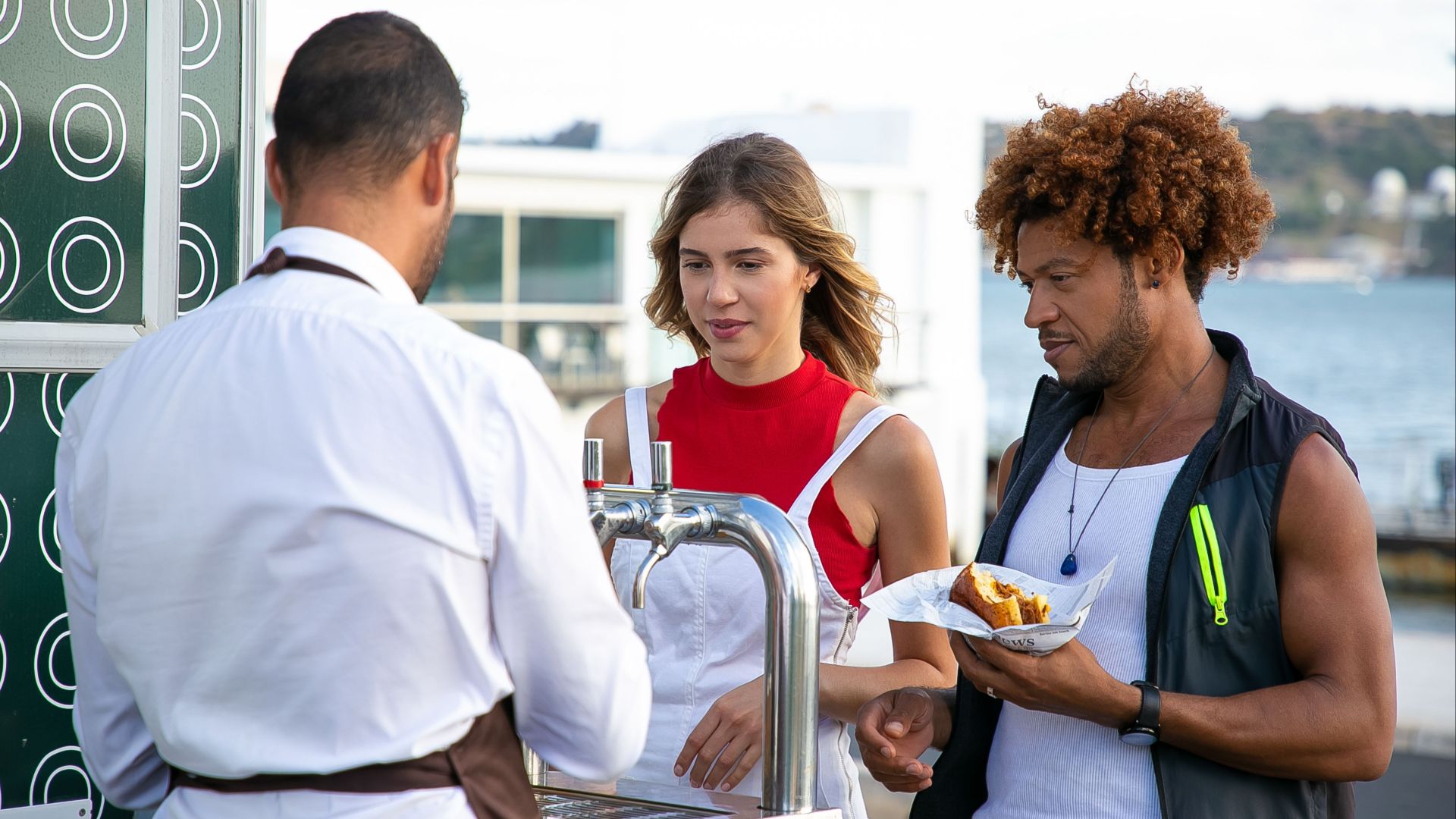 Back view of unrecognizable male bartender pouring draft beer for diverse young friends eating burger in road cafe located near lake