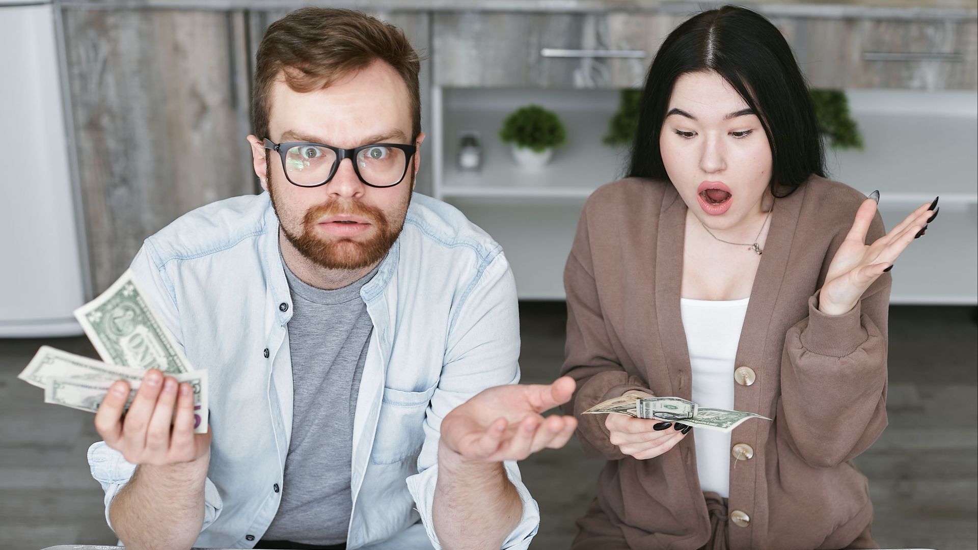 Interracial couple shocked while handling cash in a modern kitchen. Captivating expressions indoors.